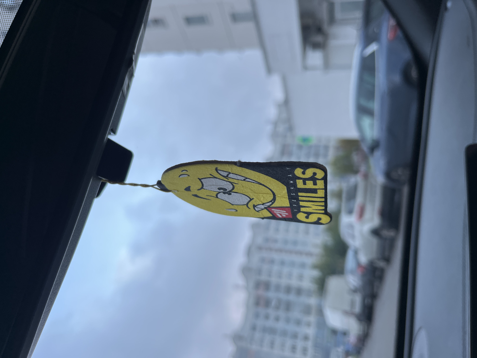 An air freshener with a cartoon smiley face and the word "SMILES" printed on it hangs from a string in the foreground, obscuring a portion of the view through the car's windshield. The air freshener is yellow with black lettering and a red and black logo. In the background, several cars are parked in a lot, and beyond them, tall residential buildings are visible under a cloudy sky. The scene appears to be in an urban environment, likely a city, possibly Minsk, Belarus, given the context of the request. The lighting suggests it is daytime.