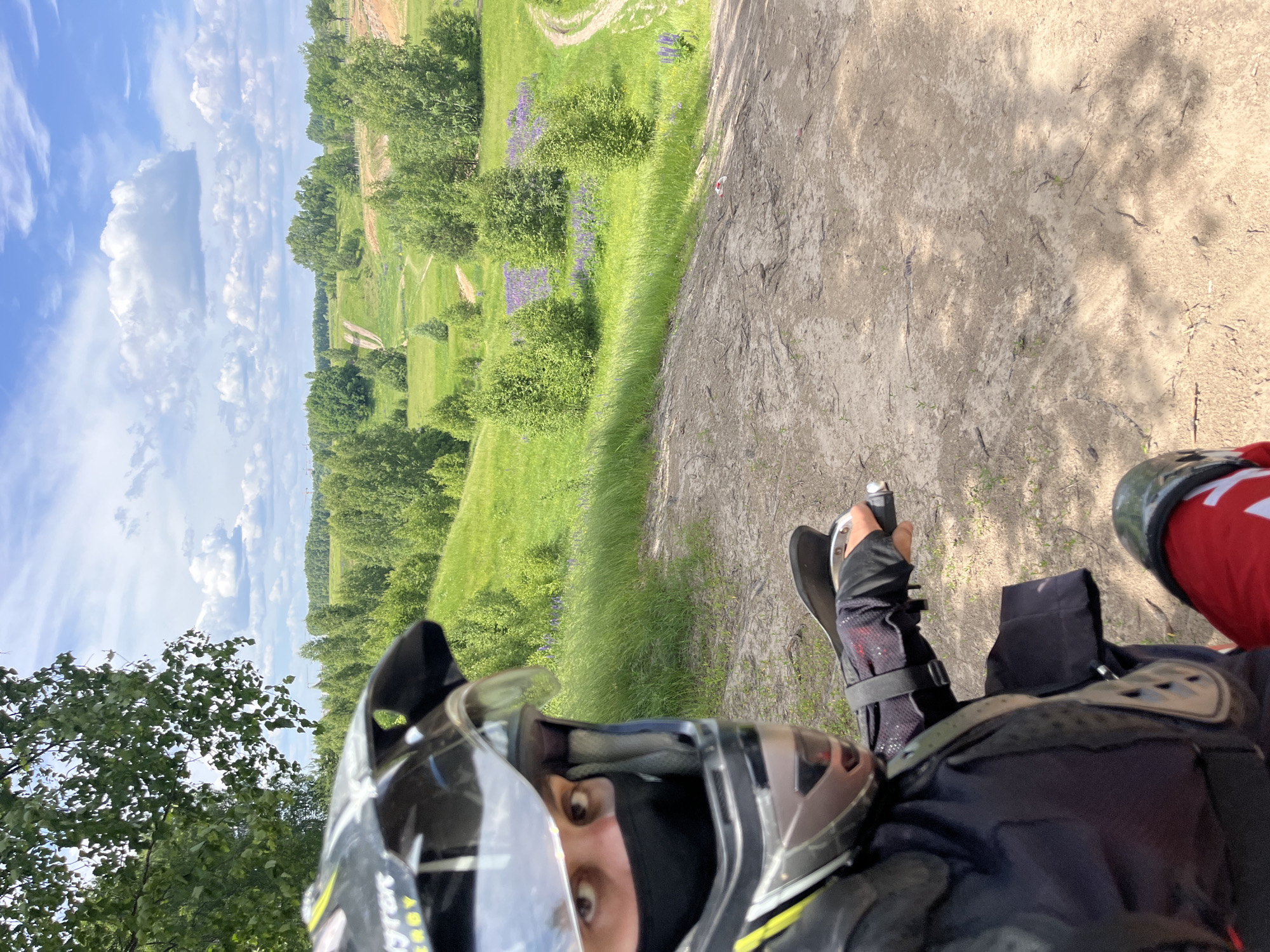 A person wearing a motocross helmet, black mask, and protective gear is on an all-terrain vehicle (ATV). The ATV is on a dirt path that slopes downwards. In the background, there is a lush green landscape with rolling hills, trees, and patches of purple flowers under a blue sky with scattered clouds. The scene appears to be an outdoor recreational area, possibly a motocross track, located in or near the city of Novochuvashsky, Russia. The individual's right hand is visible on the ATV's handlebars.