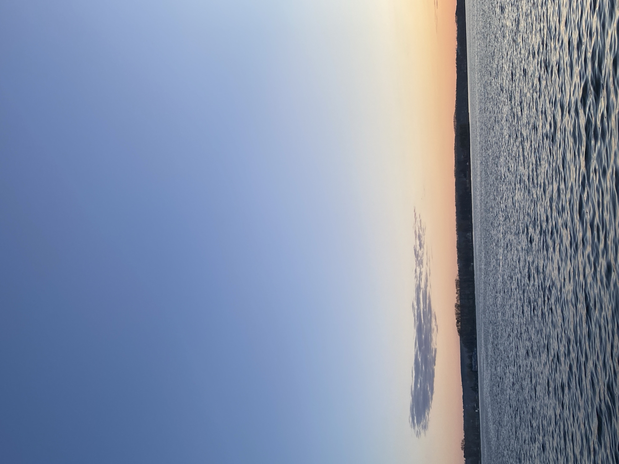 The image depicts a body of water next to a shoreline under a sky that transitions from blue to a peachy-orange color. Ripples are visible on the water surface. A narrow strip of land separates the water from the sky, with trees and possibly buildings. A cloud is present. The location may be Votkinsk, Russia.