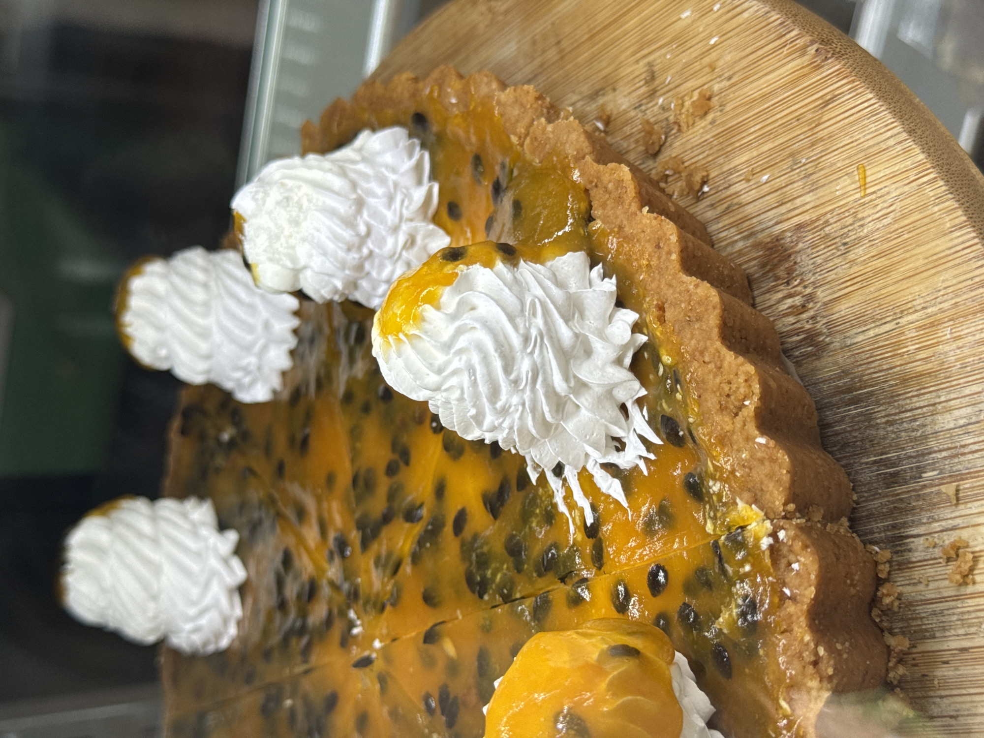 A close-up, eye-level shot shows a passion fruit tart on a light-colored wooden serving board. The tart is round with a crimped crust and is filled with bright orange passion fruit pulp studded with black seeds. Four swirls of white whipped cream are artfully placed on top of the tart, with one partially obscured by the tart's edge. A slice has been cut and removed from the tart on the left side. The background is blurred, showing a dark, indistinct environment. The scene appears to be a food display, possibly in a cafe or restaurant in Kuta Selatan, Indonesia.