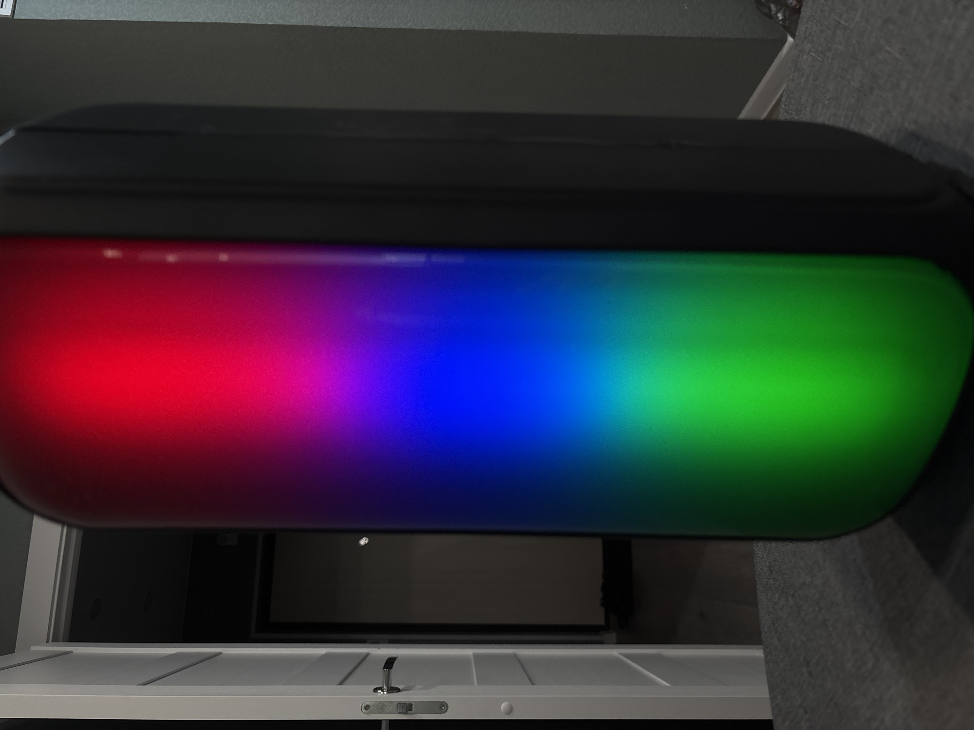 A wide-angle, horizontal shot shows a black, oval-shaped object with a colorful LED light strip running across its face. The light strip displays a gradient of colors from left to right, starting with red, transitioning through purple and blue, and ending with green. The object is positioned horizontally, appearing to be mounted or placed on a surface. Below the object, a portion of a white door with horizontal panels is visible, featuring a silver door handle and lock mechanism. The background behind the object is dark and appears to be an interior space, possibly a room with dark walls and a dimly lit area with a screen or dark surface. The lighting in the scene is dominated by the bright LED strip on the object. There is no visible text in the image. The overall impression is of a modern electronic device with customizable lighting.