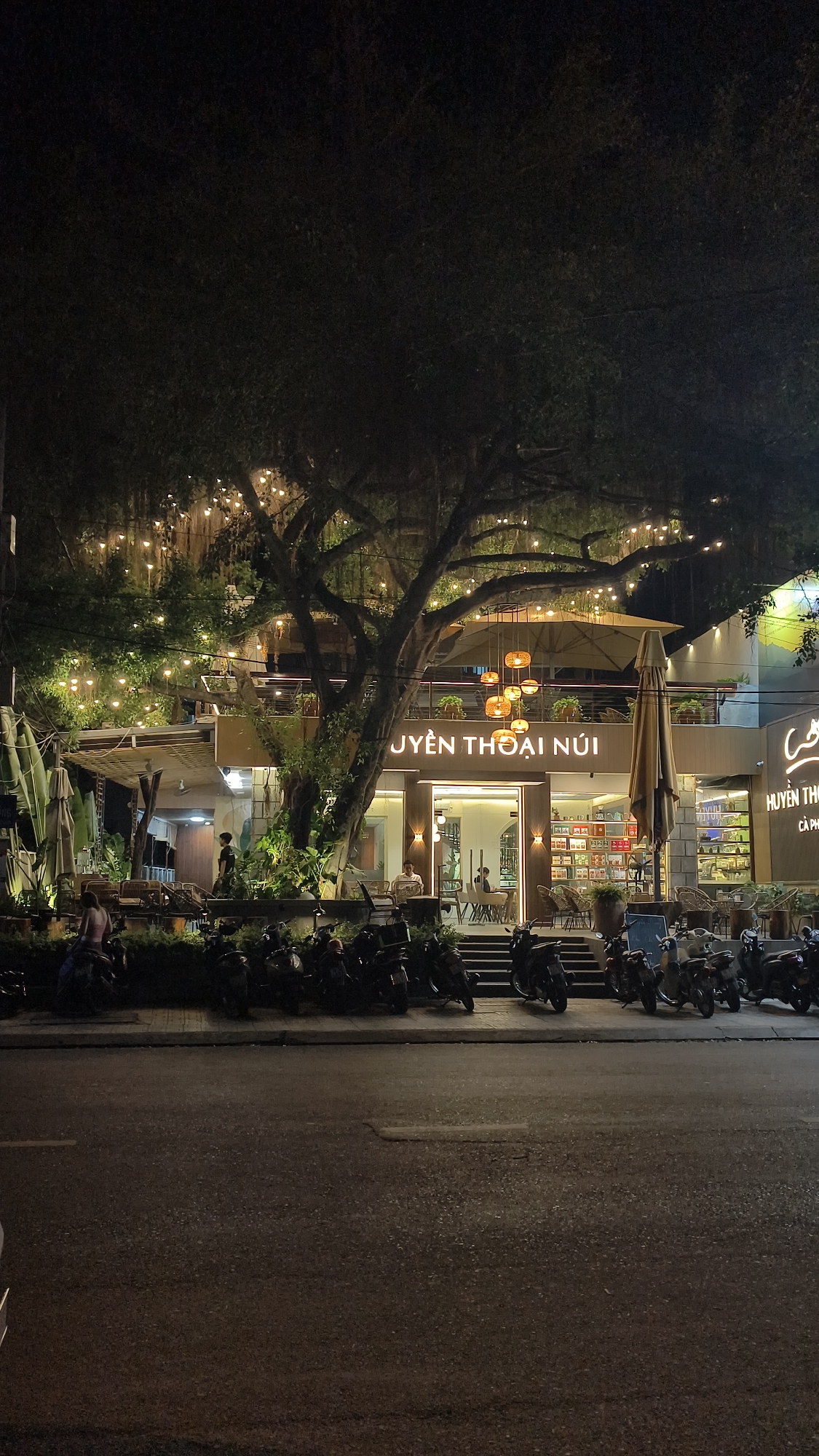 An exterior nighttime street-level shot captures a well-lit, multi-story building with a prominent tree in front. The building appears to be a commercial establishment, possibly a restaurant or cafe, with large glass windows revealing interior seating and decor. Numerous string lights are draped across the tree's branches and illuminated, creating a festive atmosphere. A large, tan umbrella is positioned outside the entrance, and several tables and chairs are visible on an outdoor patio area. In front of the building, a row of parked motorcycles is lined up along the sidewalk. The street in the foreground is paved asphalt, with a visible white lane marking. The overall scene suggests a lively urban setting at night, likely in a Southeast Asian city given the style of motorcycles and the building's architecture.