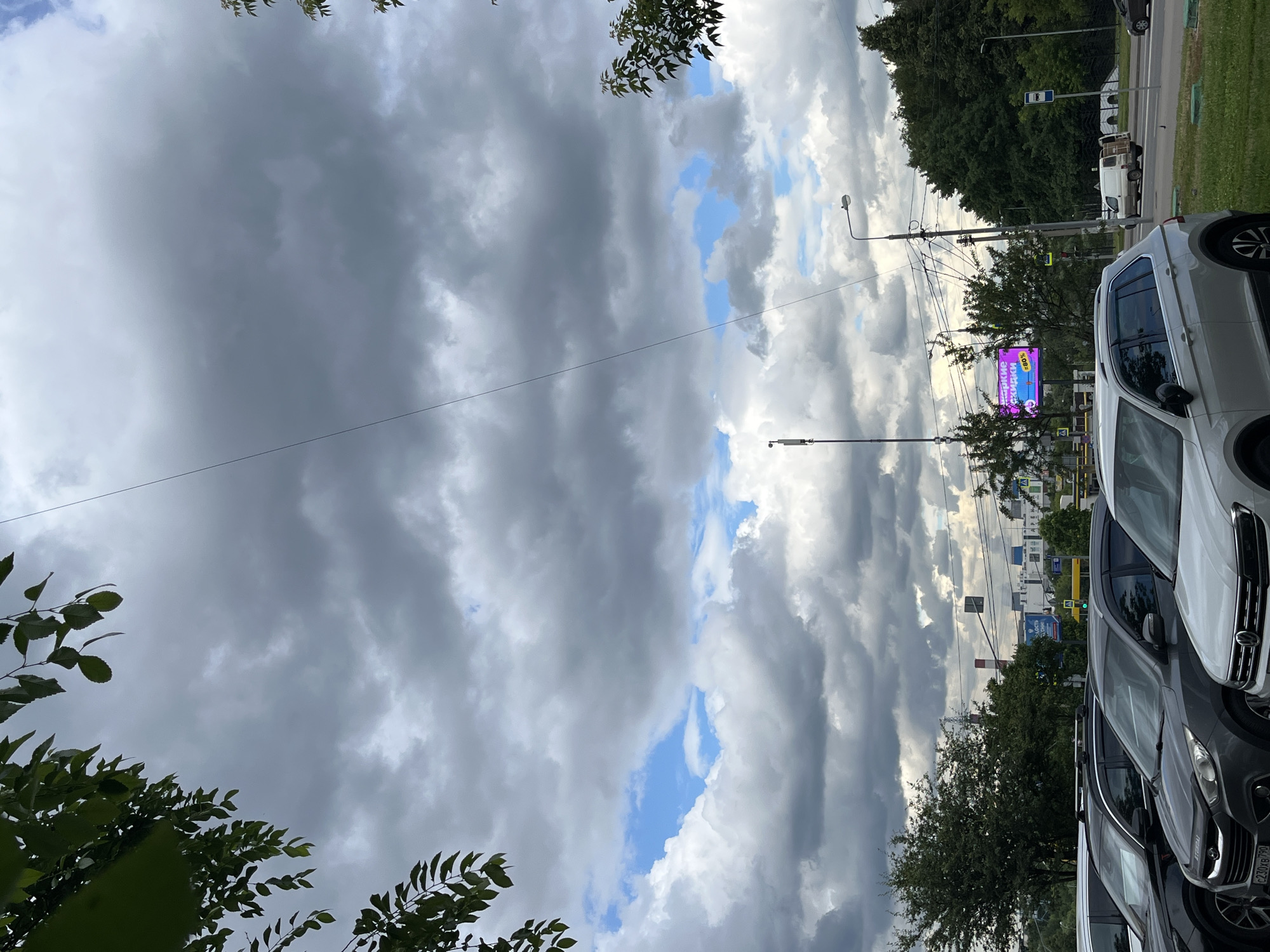 The image depicts a street scene in Moscow, Russia, with a focus on a gray, cloudy sky. Dense, dark clouds dominate the upper portion of the frame, interspersed with patches of blue sky. Sunlight is diffused by the clouds, suggesting either an overcast day or early morning/late afternoon.
In the foreground and midground, several cars are parked along the side of a road. These include a white SUV on the right, a dark gray SUV in the center, and other vehicles partially visible behind them. The cars appear to be modern models. A bright purple billboard with text is visible behind the dark gray SUV, positioned in front of trees. The billboard displays text that reads "ЯРКИЕ СКИДКИ" and "80%".
Trees with green foliage line the road and a grassy area is present on the right side of the frame. Streetlights and utility poles with numerous wires are also present, indicating an urban or suburban setting. In the background, buildings and more trees can be seen, along with glimpses of other vehicles on the road, including a white van. The overall impression is of a typical day in a Russian city, with changeable weather and everyday activities. There are no people clearly visible in the image.