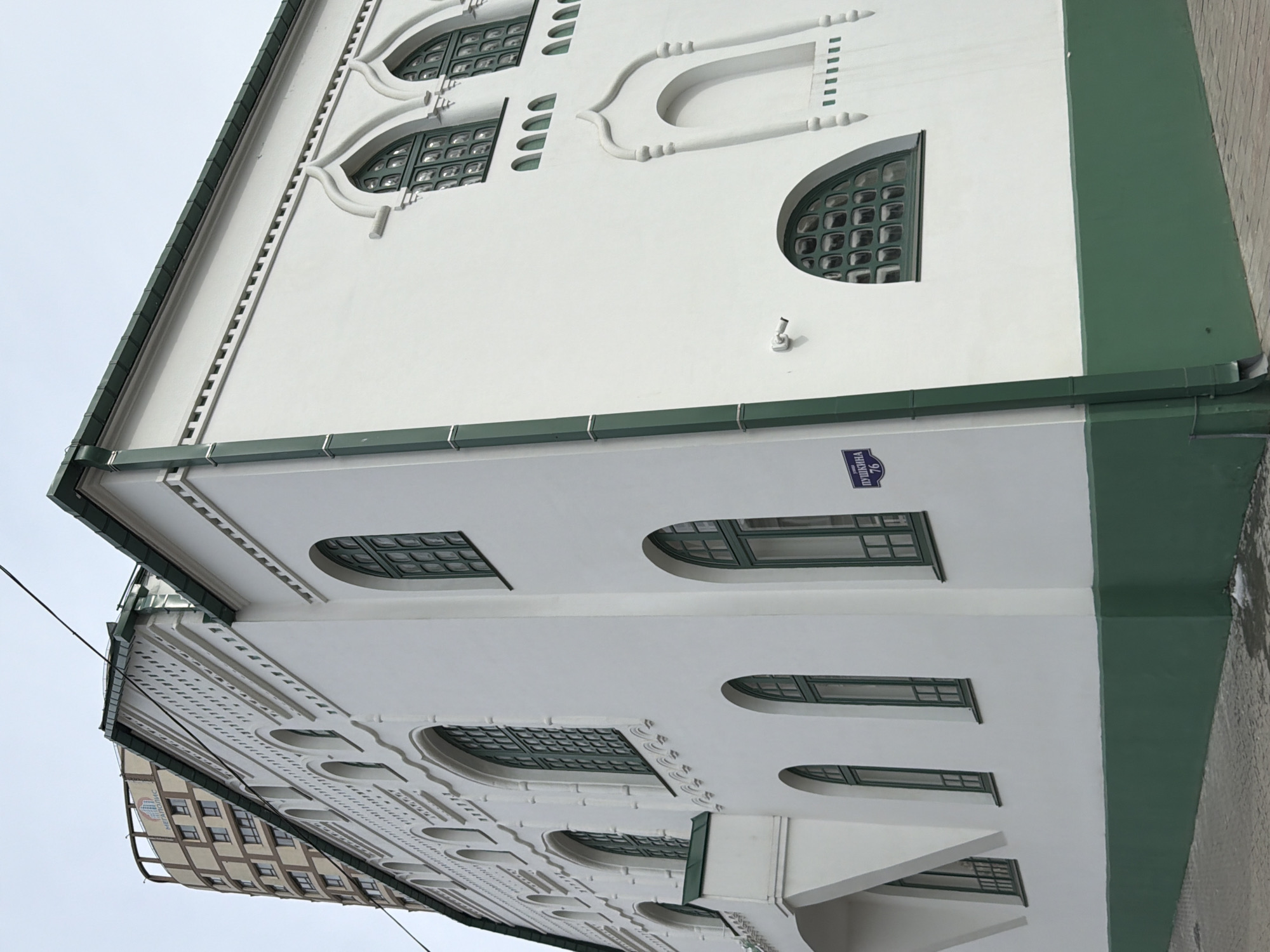 The image shows a white building with green trim in Perm, Russia. The building has a street address sign that says "Пушкина 76" (Pushkina 76). The building's facade features several arched windows with green frames and decorative white elements. The overall architectural style appears historical.