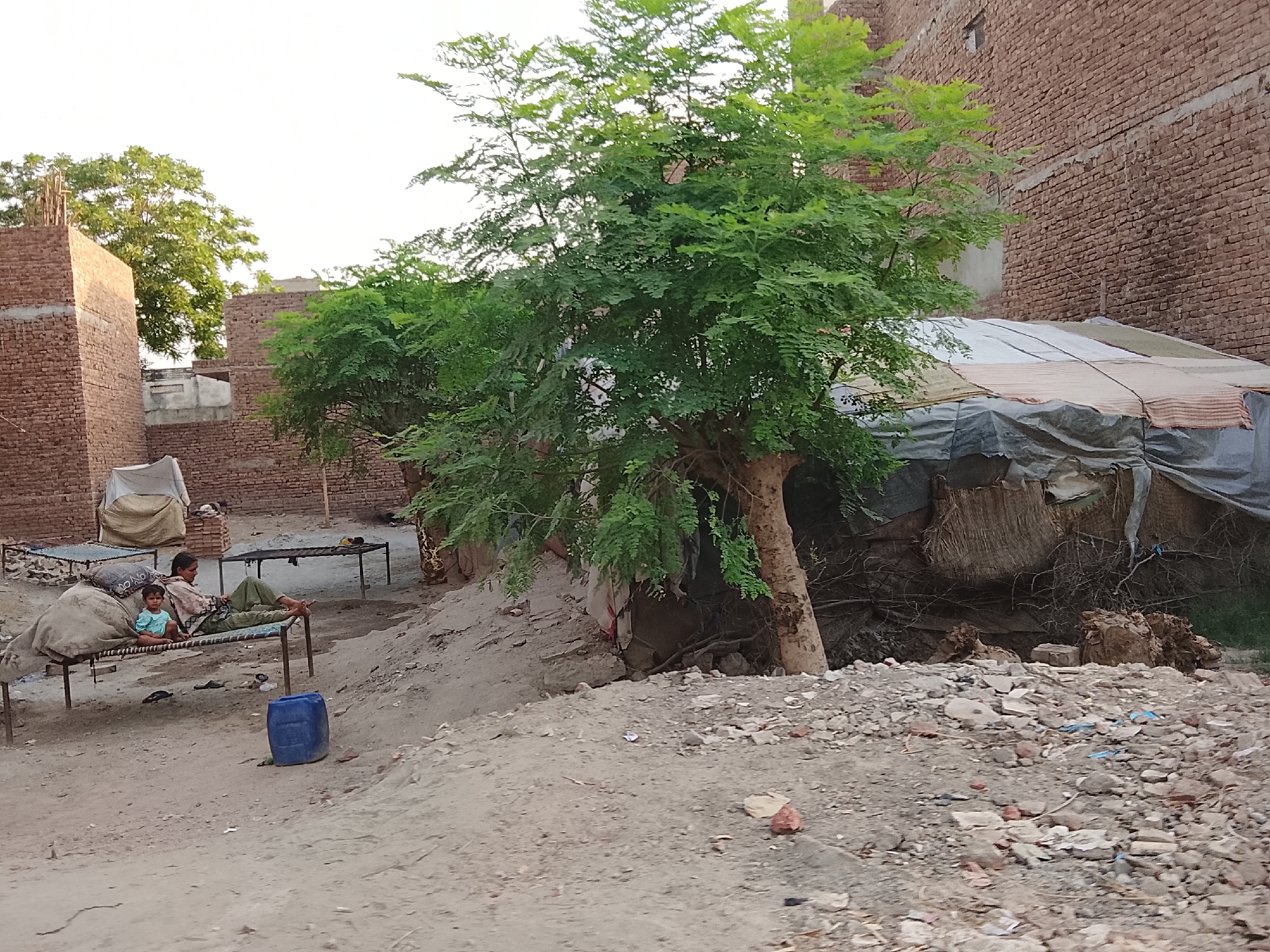A scene from an unknown area, possibly a village or impoverished area. A woman and child are sitting on a woven bed, with another empty woven bed nearby. Behind them are brick buildings, one appearing unfinished. A large green tree dominates the foreground, partially obscuring a makeshift shelter constructed from tarps and natural materials. The ground is dusty and scattered with debris. A blue plastic jug sits near the bed where the woman and child are.