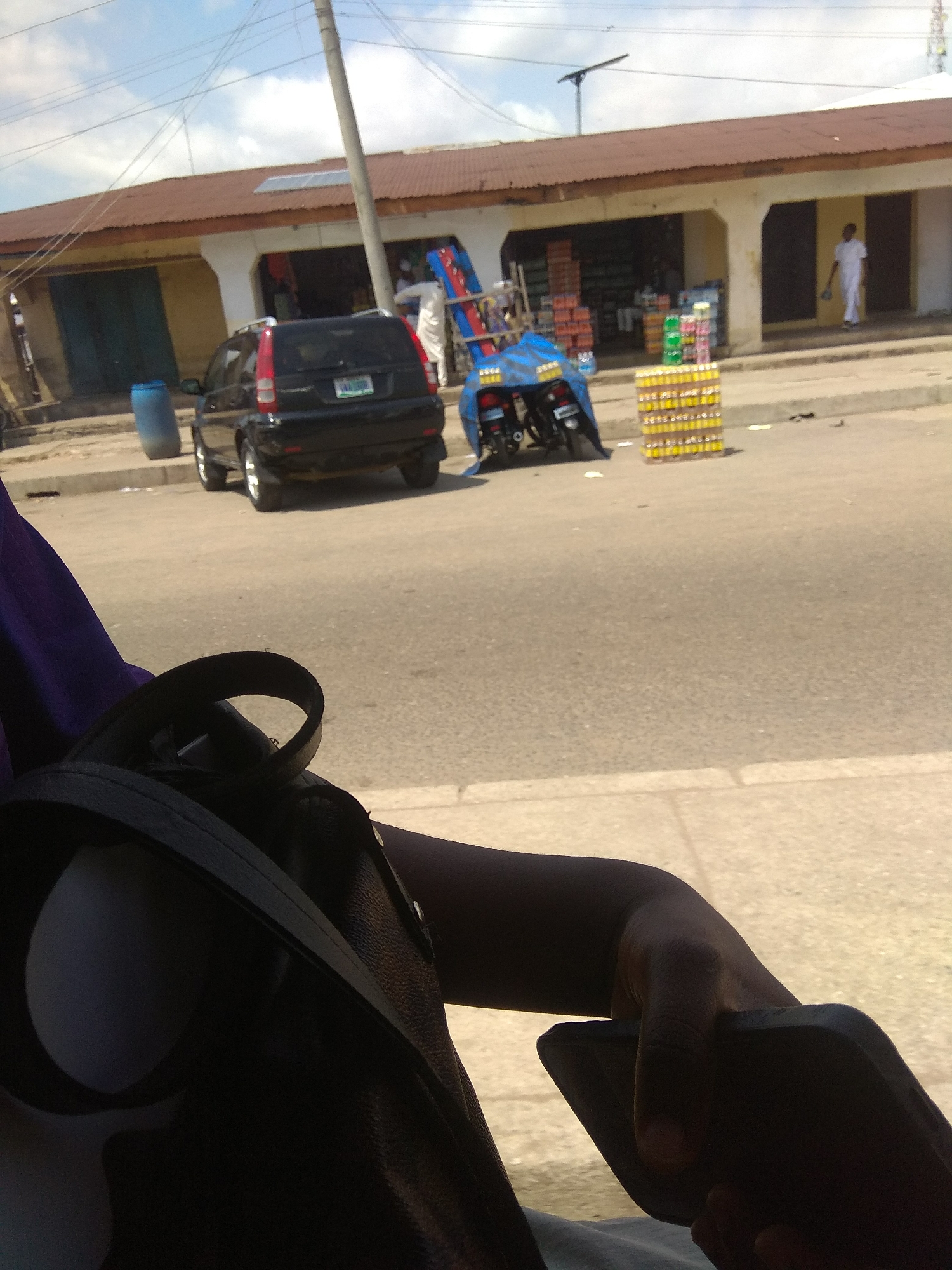 In Jalingo, Nigeria, a street scene unfolds with a dark SUV parked on the right side of a paved road. The SUV has a license plate that is partially obscured by glare. To the left of the SUV, a blue barrel stands on the side of the road. In the background, a building with a corrugated metal roof stretches across the scene. The building has open storefronts with displays of goods, including stacks of colorful crates and bottles. A person in a white garment is visible near one of the storefronts. On the left side of the image, a motorcycle covered in a blue tarp is parked. In the foreground, a person's hand holds a smartphone, and a dark purse or bag is visible. The sky above is cloudy.