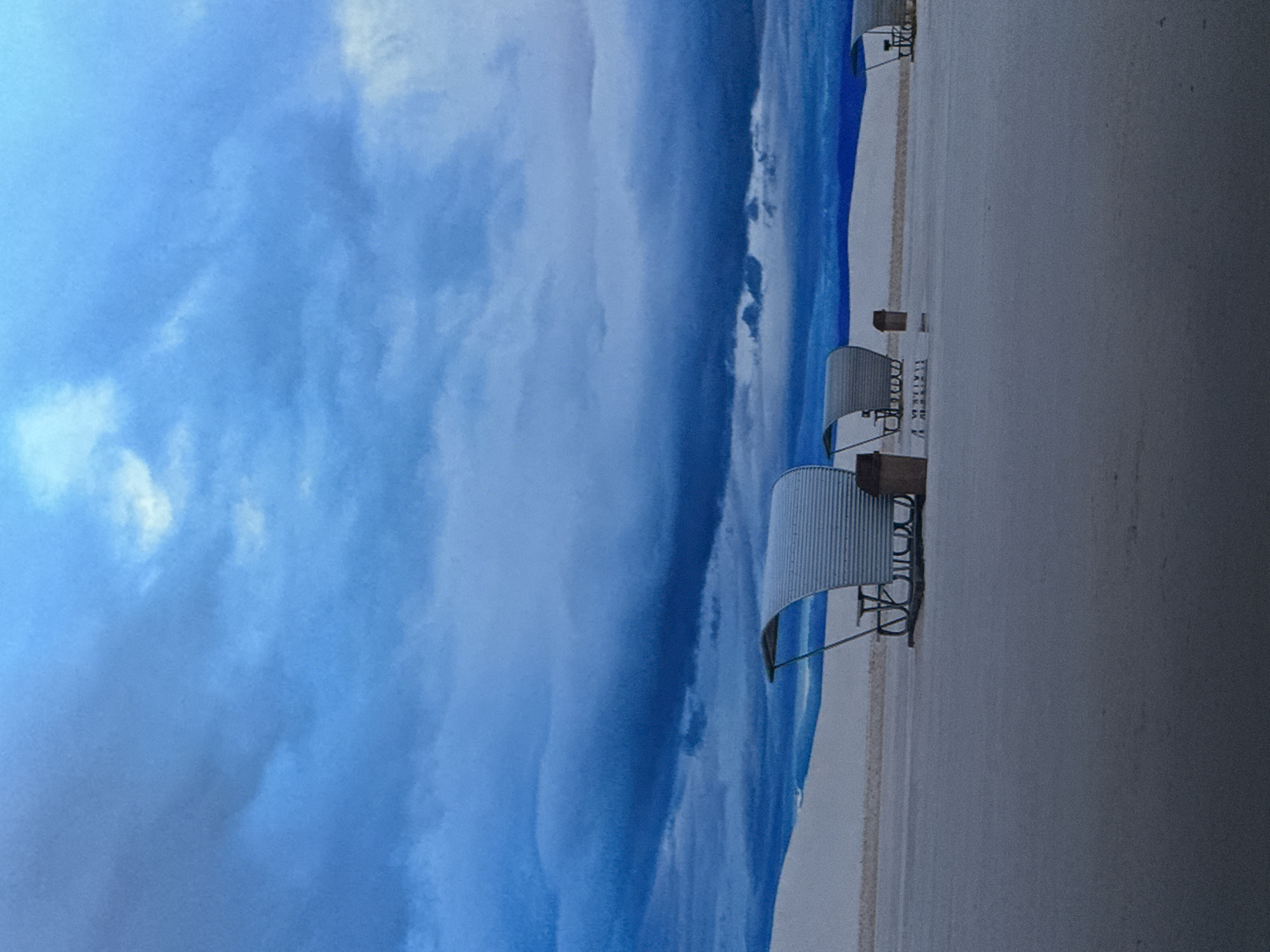 This is a horizontal, low-angle outdoor photograph taken during dusk or early morning at White Sands National Park in New Mexico. The image is dominated by a dramatic sky filled with dark, bruised blue and grey storm clouds that are illuminated by a faint lighter blue and white near the horizon. Below the sky, smooth, undulating white sand dunes stretch horizontally across the midground. In the foreground, the flat, pale sand appears almost reflective. Scattered across the midground are several ribbed, metal picnic shelters with attached benches and tables, designed to resemble covered wagons. One shelter is closer to the left side of the frame, while others are further in the distance. There are no people visible in the image. The overall atmosphere is serene and somewhat ominous due to the stormy sky.