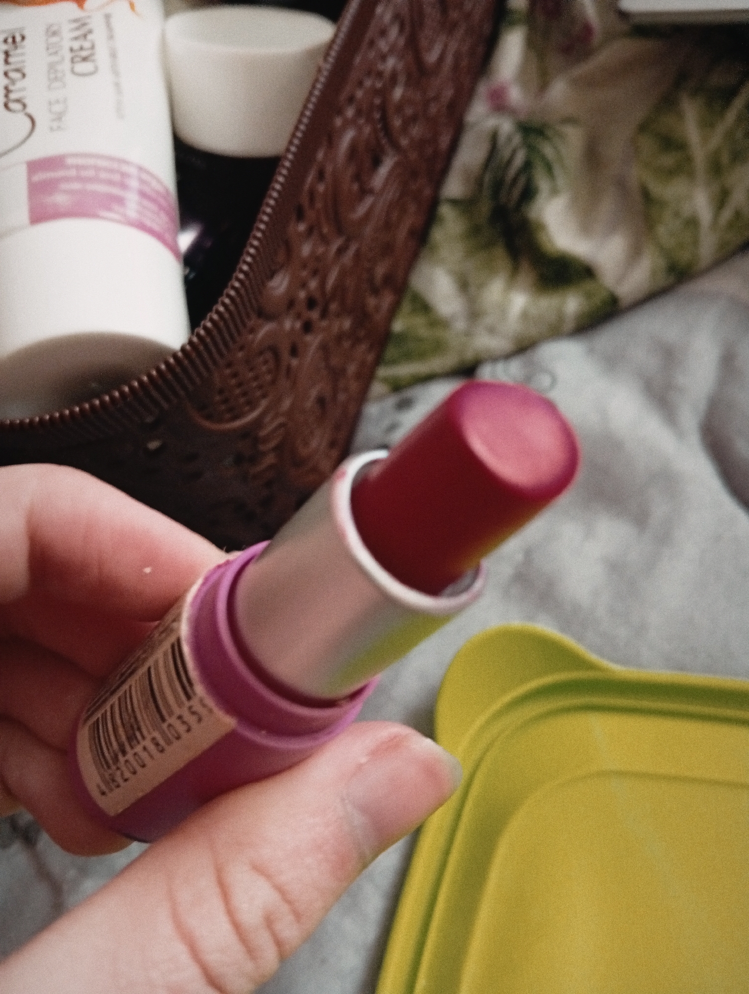 A human hand holds a partially opened tube of lipstick. The lipstick's color is a deep reddish-pink. The tube is silver with a pink bottom section and has a barcode. The background contains a brown wicker basket filled with items, including a white tube labeled "Caramel FACE DEPILATORY CREAM," and a white jar. A green and white patterned fabric and a gray surface are also visible. A yellow plastic tray is in the lower right of the frame. The location is not determinable from the given image.