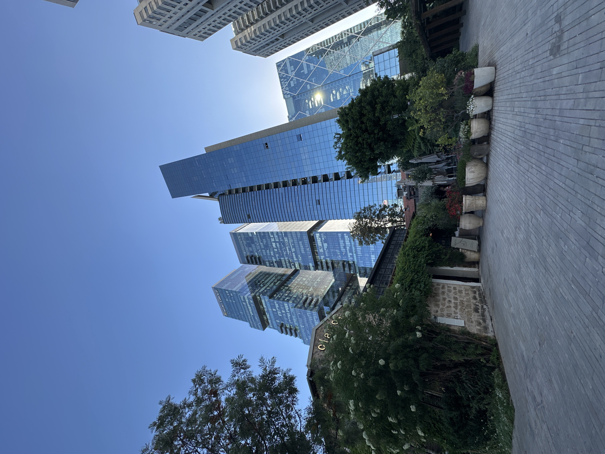 The image captures a view in Tel Aviv, Israel, showcasing a mix of modern architecture and green spaces. Several skyscrapers dominate the skyline, constructed with reflective glass facades that mirror the clear blue sky. One building is labeled as "Hagag Group." Lush trees and flowering plants create a green environment at ground level, juxtaposing the urban landscape. Along a brick walkway, a series of large, round, antique-style vases hold plants. A stone building with climbing vegetation is also visible. Overall, the scene presents a modern city environment integrated with natural elements.