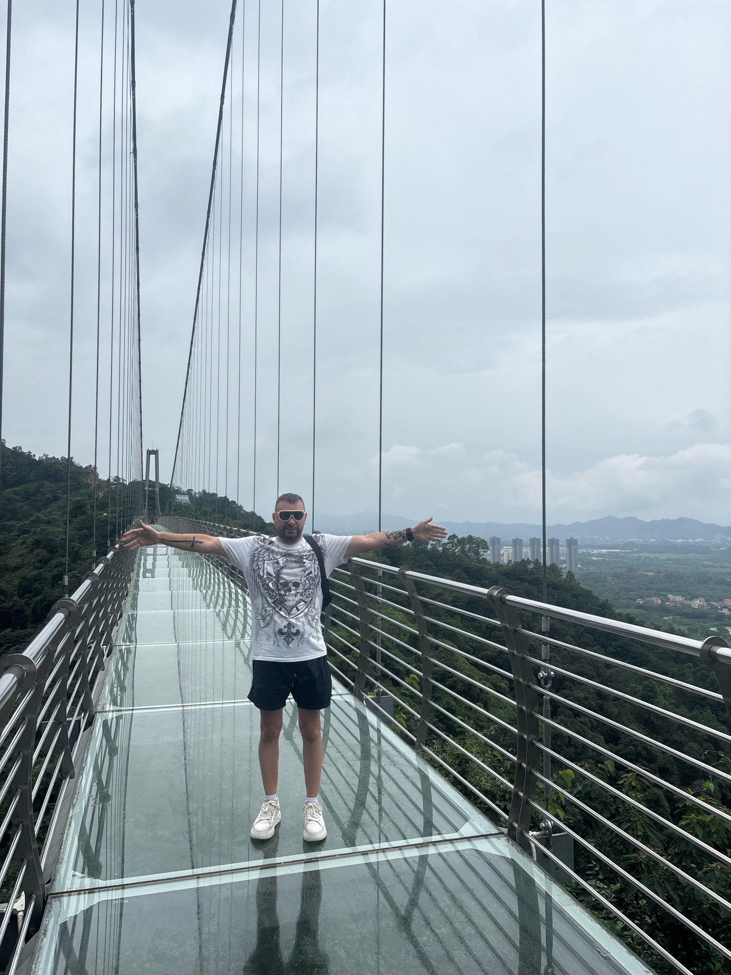 A man stands on a glass-bottomed suspension bridge, his arms outstretched. He is wearing a white t-shirt with a skull graphic, black shorts, and white sneakers. He has sunglasses on and a backpack. The bridge is surrounded by lush green mountains and a hazy cityscape can be seen in the distance. The bridge is part of the Foshan Baili Bridge, a tourist attraction in Foshan, China. The sky is overcast, and the glass walkway reflects the surrounding environment.