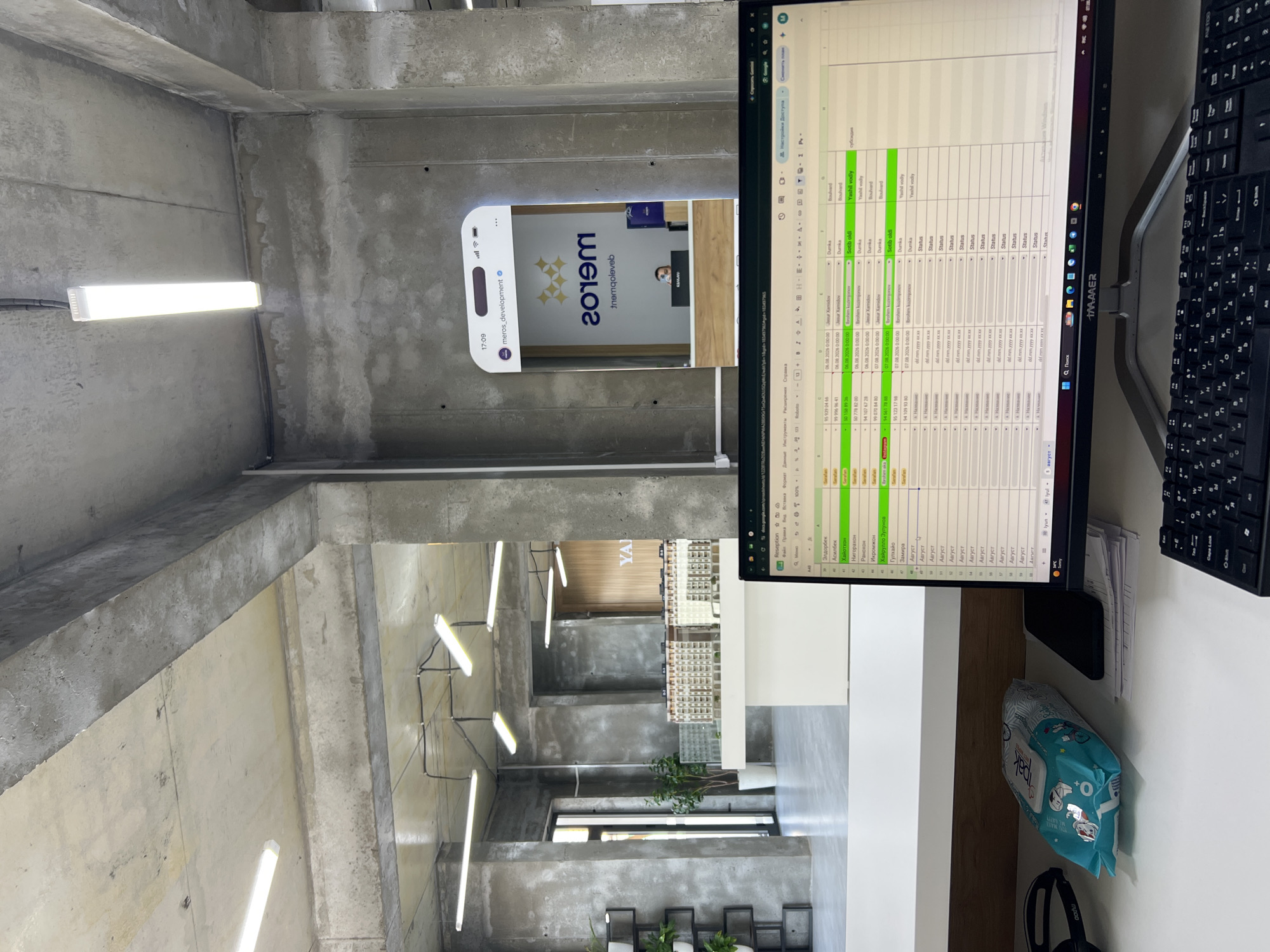 The image depicts an office interior with a modern, industrial aesthetic. Concrete walls and ceilings with exposed beams and conduits are prominent. The scene is viewed from the perspective of someone sitting at a desk, looking towards a reception area in the background.
In the foreground, a computer monitor displays a spreadsheet, likely for office management or data entry. The spreadsheet has columns for names, dates, and other information, with some rows highlighted in green. To the left of the monitor, a packet of wet wipes and a smartphone are visible on the desk.
The background reveals a reception area with a counter and a logo on the wall that reads "meross". A person is seated behind the counter, partially visible. A large, reflective surface, possibly a mirror or a large screen, displays a social media interface with the username "meros_development". There are also architectural models of buildings placed on a white counter in the middle ground.
The overall impression is of a contemporary workspace, likely located in a business setting within the city of Shahrixon, Uzbekistan, given the context of the reception area's signage and potential local business operations. The lighting is primarily from fluorescent tubes mounted on the ceiling and possibly natural light entering from unseen windows. The environment appears clean and organized.