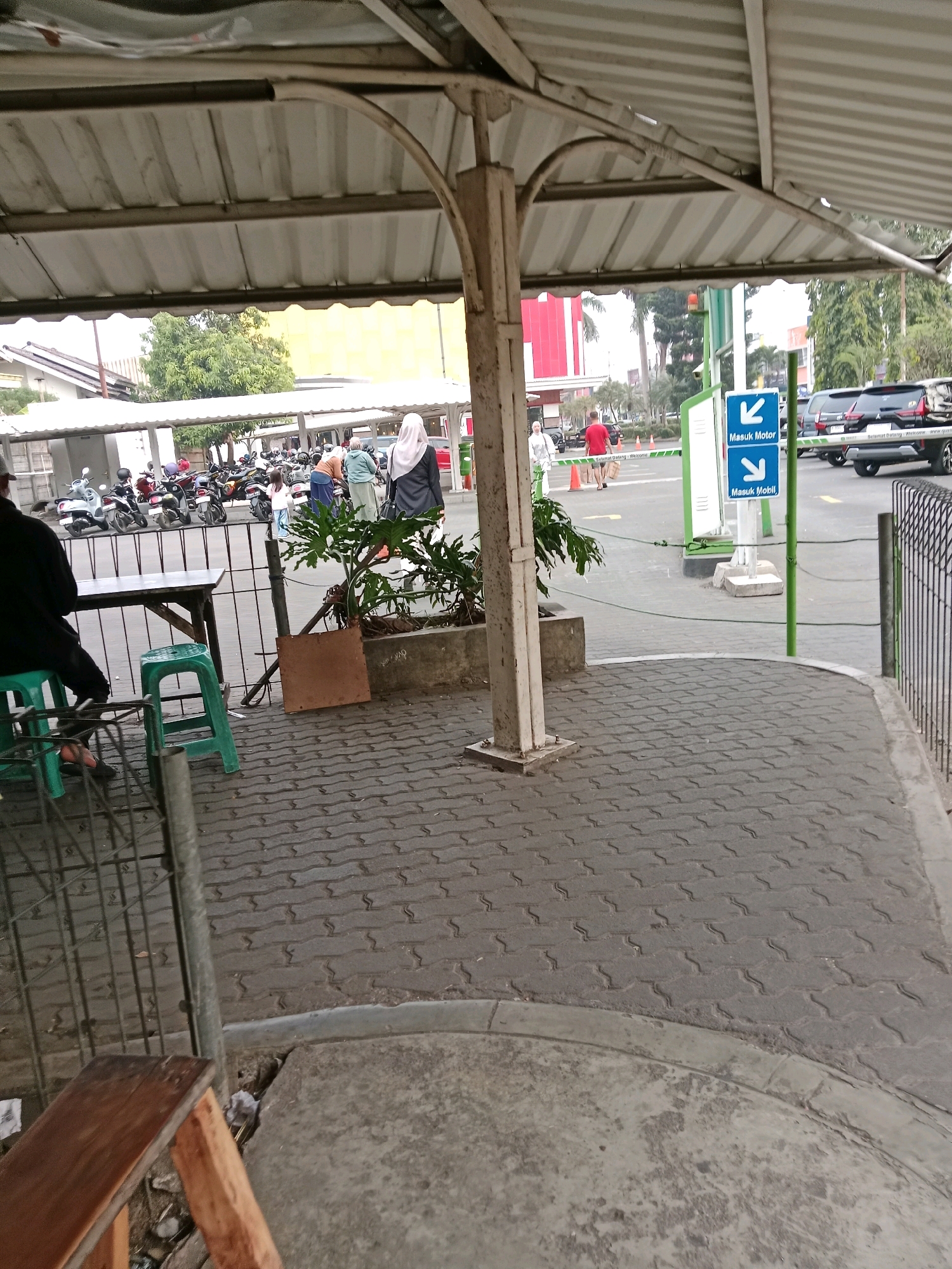 The image shows a covered outdoor area, likely a parking lot entrance, in Cianjur, Indonesia. A person in a black shirt is seated on a green stool to the left, with a table in front of them. In the background, several motorcycles are parked under a white awning. To the right, two blue signs with white arrows indicate "Masuk Motor" (Motorcycle Entrance) and "Masuk Mobil" (Car Entrance). A black SUV and other vehicles are visible in the background. Several people are walking in the distance, some wearing headscarves. The ground is paved with interlocking gray bricks. The environment suggests a public space with regulated vehicle entry.
