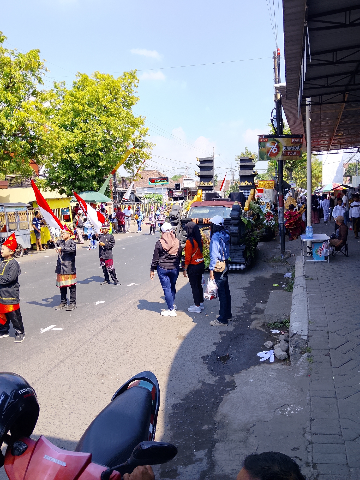 This image captures a street scene in Kertosono, Indonesia, likely during a procession or parade. The foreground features a red scooter with a black helmet. In the mid-ground, several individuals are participating in what appears to be a cultural event. Two young boys in traditional attire, one holding a red and white flag, are prominently positioned. Other participants, some in casual clothing, are also visible. To the right, a sidewalk lined with shops and a few people, one seated at a small table, is present. Further down the street, a decorated float or vehicle is visible, adorned with dark structures and floral arrangements. Trees and buildings line the background, under a bright blue sky with scattered clouds. The street is paved, with some wet patches and debris on the side. A sign with "76" and "MANGGA" is displayed on a pole.