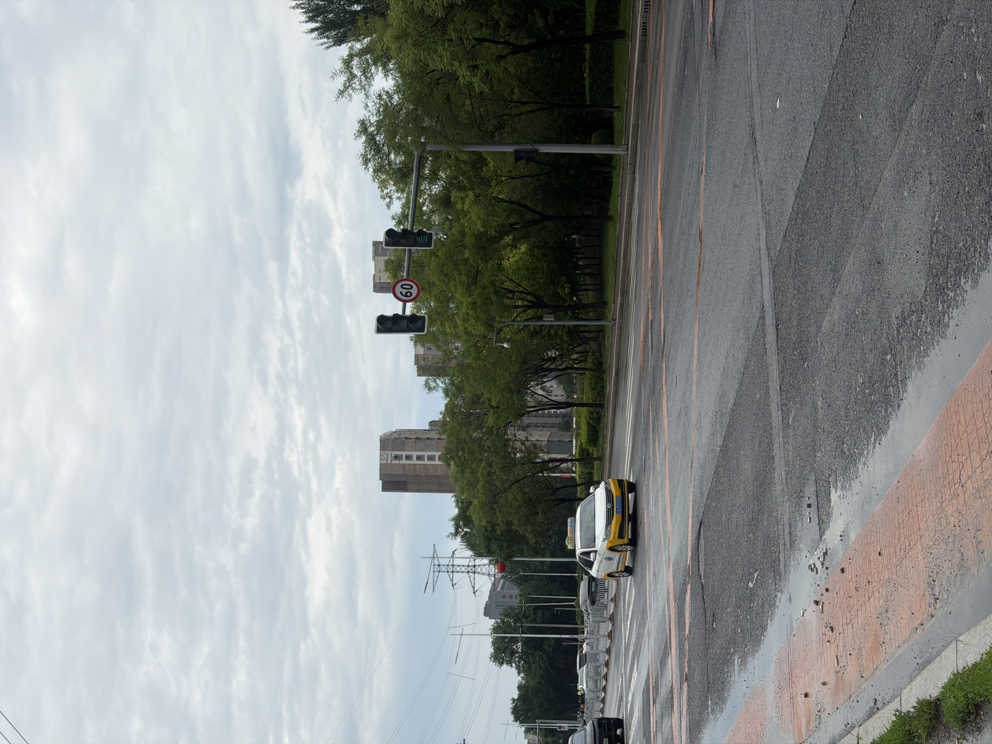 The image depicts a cloudy day on a street in Shenyang City, China. A yellow and white taxi is driving in the right lane of the road. The road is wet, suggesting recent rain, and has orange markings indicating pedestrian crossings. Traffic lights are visible, with the right-side light showing green. Buildings and trees line the street. In the background, there are tall apartment buildings and power transmission towers.