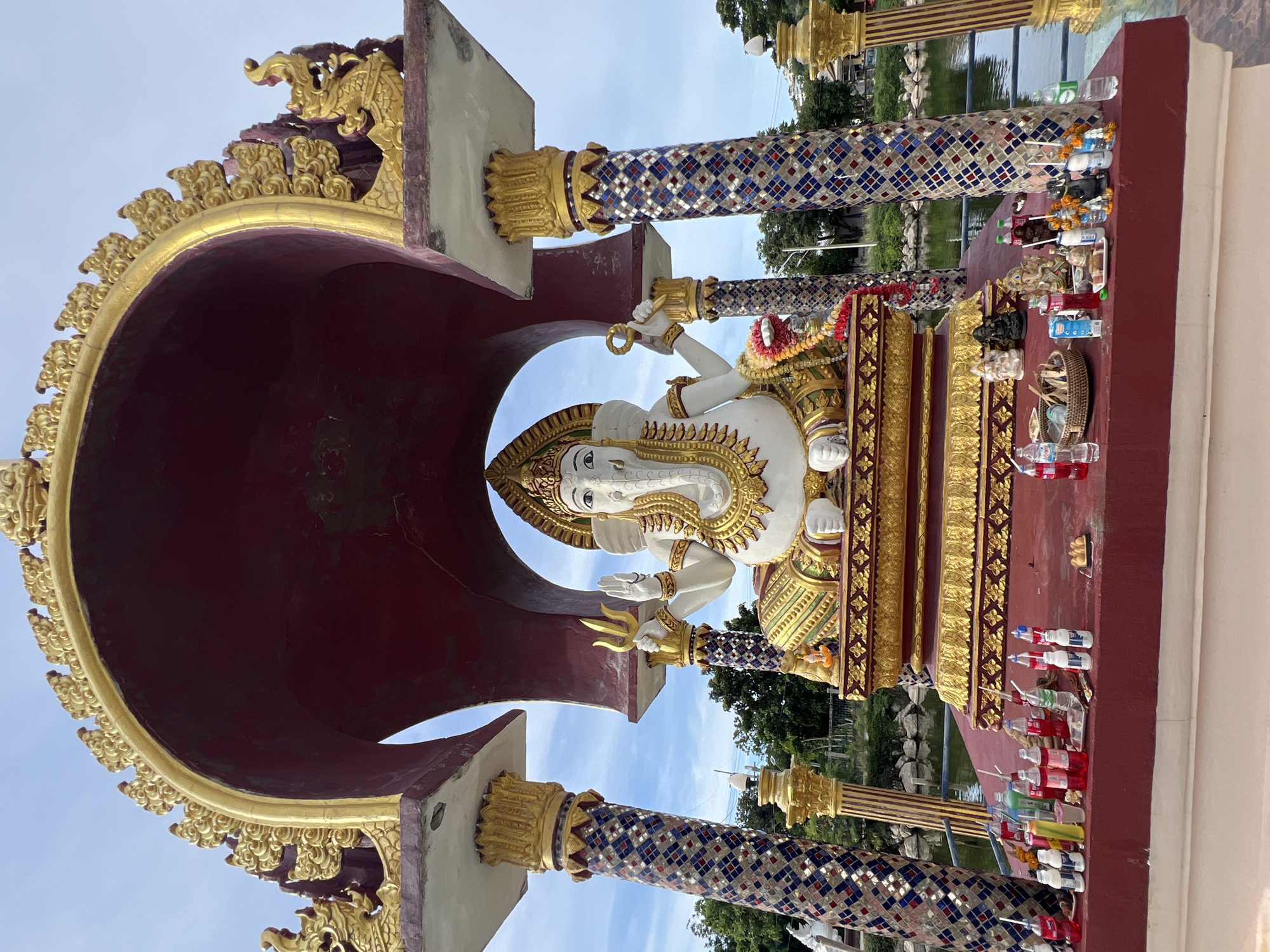 A large, white statue of Ganesha, the Hindu deity, sits on a golden throne under a red and gold archway. The statue is adorned with a gold necklace and a garland of red and white flowers. In front of the statue, on a red platform, are various offerings, including bottles of red and clear liquid, a woven basket, and small statues of deities. The scene is set outdoors, with a body of water and trees visible in the background. The sky is partly cloudy. This is the Ganesha temple at Baan Plai Laem, Koh Samui, Thailand.