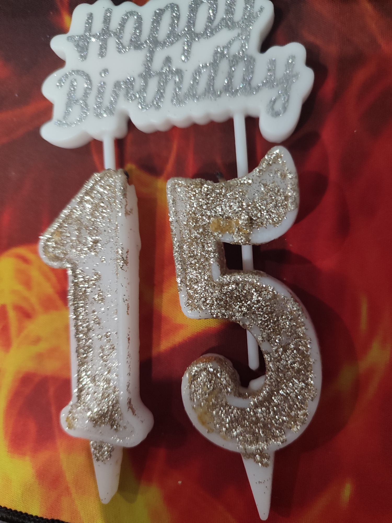 The image shows a "Happy Birthday" cake topper with two number candles, "1" and "5", forming "15". The topper and candles are white with silver glitter. The background is a red and yellow abstract pattern. The scene is indoors. There are no people or actions visible. The location in Mykolaiv, Ukraine cannot be determined from the image.