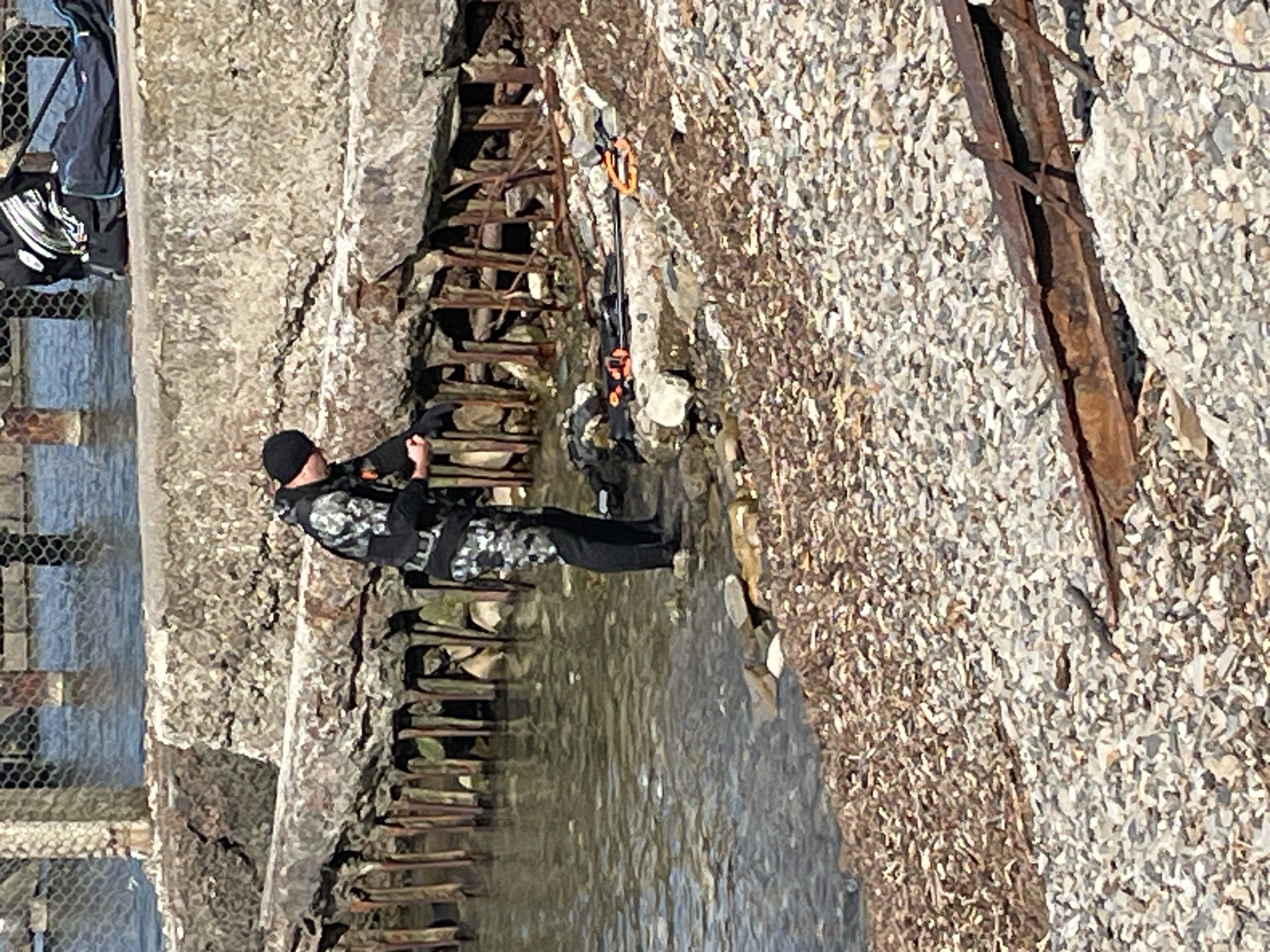 The image shows a person in a diving suit standing by the water's edge, appearing to be preparing dive gear. The scene takes place on a rocky shore with what looks like a weathered concrete structure and pilings in the background. There is also scuba diving equipment visible on the ground near the water. The location is identified as Sochi, Russia.