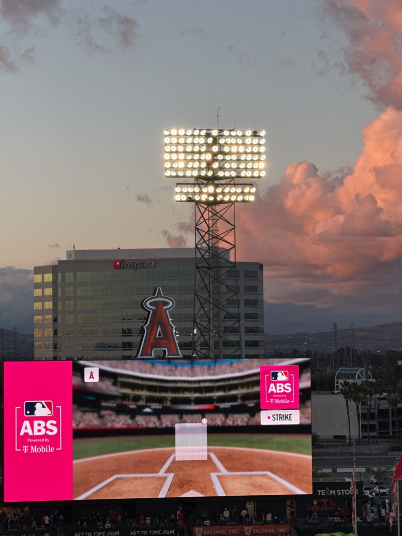 This image captures an atmospheric scene at a baseball stadium during dusk or twilight. The sky transitions from a soft blue-grey to warm pink and orange hues with scattered clouds, indicating the setting sun.
In the foreground, a large, brightly lit video scoreboard dominates the lower half of the frame. It displays a graphic of a baseball field from behind home plate, with a transparent strike zone box and a small baseball visible. The scoreboard features prominent branding: on the left, a magenta panel shows "ABS" with a baseball player logo, "Powered by T Mobile," and a small "A" logo. A similar magenta panel on the right displays "ABS," "T Mobile," and "• STRIKE," also with a small "A" logo.
Above the scoreboard, a tall light tower with numerous rows of brilliant white stadium lights illuminates the scene. Behind the light tower and the scoreboard is a large, modern building with a prominent red "A" logo with wings, characteristic of the Los Angeles Angels baseball team. The building itself has "Impact" visible as signage.
Below the scoreboard, smaller text advertisements are visible on a barrier, reading "NITTO TIRE.COM" (repeated) and "SALINAS, INC." (repeated). Further to the right, a "TEAM STORE" sign can be seen. In the distance, rolling hills or mountains are silhouetted against the fading light. The overall scene suggests the beginning or end of a baseball game, set against a picturesque sunset at Angel Stadium in Anaheim, United States.