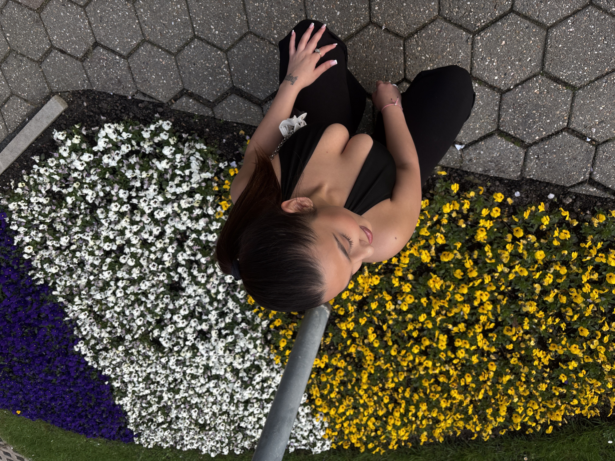 A high-angle shot captures a woman seated on a paved area next to a flowerbed. The pavement is constructed of gray hexagonal blocks. The flowerbed is filled with pansies in sections of purple, white, and yellow. A gray pole bisects the flowerbed. The woman is wearing black pants and a black top. She has dark hair and is looking down at the flowers. A small tattoo is visible on her left wrist. She is wearing a ring on her left ring finger. A white object, possibly a charm, is visible on her left shoulder. The location of the scene is most likely a park or green space within the city of Mönchengladbach, Germany.