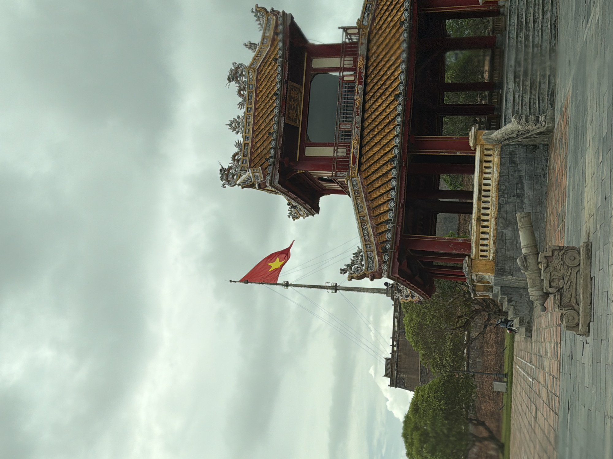 In Phú Xuân Ward, Vietnam, an outdoor scene depicts a traditional architectural structure with a red and gold tiered roof, possibly a pavilion or temple, adorned with dragon sculptures. A Vietnamese flag, red with a yellow star, flies from a tall mast to the left of the structure. In the foreground, a stone cannon rests on a tiled courtyard. Behind the pavilion, a section of a large, ancient stone wall is visible, with green trees in the mid-ground. The sky is overcast with clouds. A person on a scooter is parked near the stone wall.