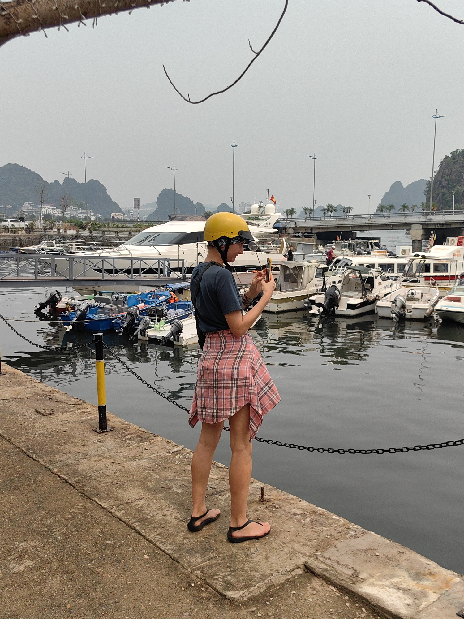 This image depicts a person standing on a concrete pier in a harbor in Hong Gai Ward, Vietnam, on an overcast day. The person, viewed from behind and slightly to the side, is wearing a yellow helmet, a dark t-shirt, a red and white plaid shirt tied around their waist, and black flip-flops. They are actively holding a smartphone, appearing to take a photograph or video of the scene.
The harbor is busy with various boats docked in the calm water. A prominent large white yacht, adorned with a Vietnamese flag at its stern, is visible in the mid-ground. Closer to the pier, several smaller blue motorboats and other white leisure boats with outboard motors are moored. Reflections of the boats and the chain barrier along the pier are cast upon the water's surface.
Behind the boats, a bridge with streetlights and palm trees spans across the water, leading to a cityscape with buildings nestled against the base of towering karst mountains. These distinctive limestone formations dominate the background under a hazy, grey sky. A tree branch with bare twigs extends into the top left corner of the frame. The concrete pier features a black chain barrier supported by black and yellow poles. Partially visible text on some boats includes "THANH 02" and "DK 42...". The scene captures a moment of observation and documentation within a scenic Vietnamese port.