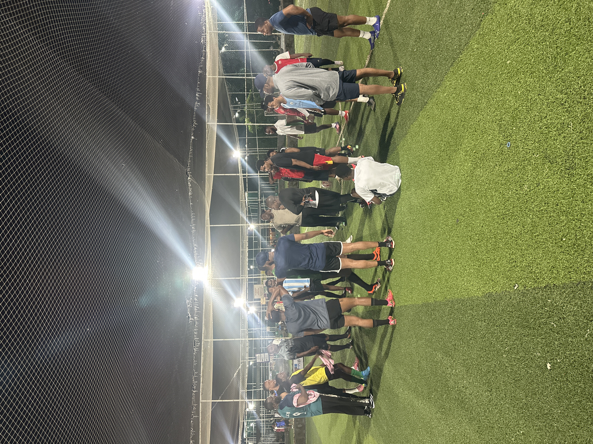 The image depicts a group of men on an artificial turf soccer field at night. The field is illuminated by overhead lights, with bright beams cutting through the darkness. A net enclosure surrounds the field, suggesting it is an indoor or covered facility. Most of the men are wearing athletic clothing, including shorts and jerseys, and appear to be engaged in conversation or a team huddle. Some are wearing soccer cleats. One individual in the foreground is seated on the ground. The setting is likely a sports facility in Abuja, Nigeria, given the context provided.