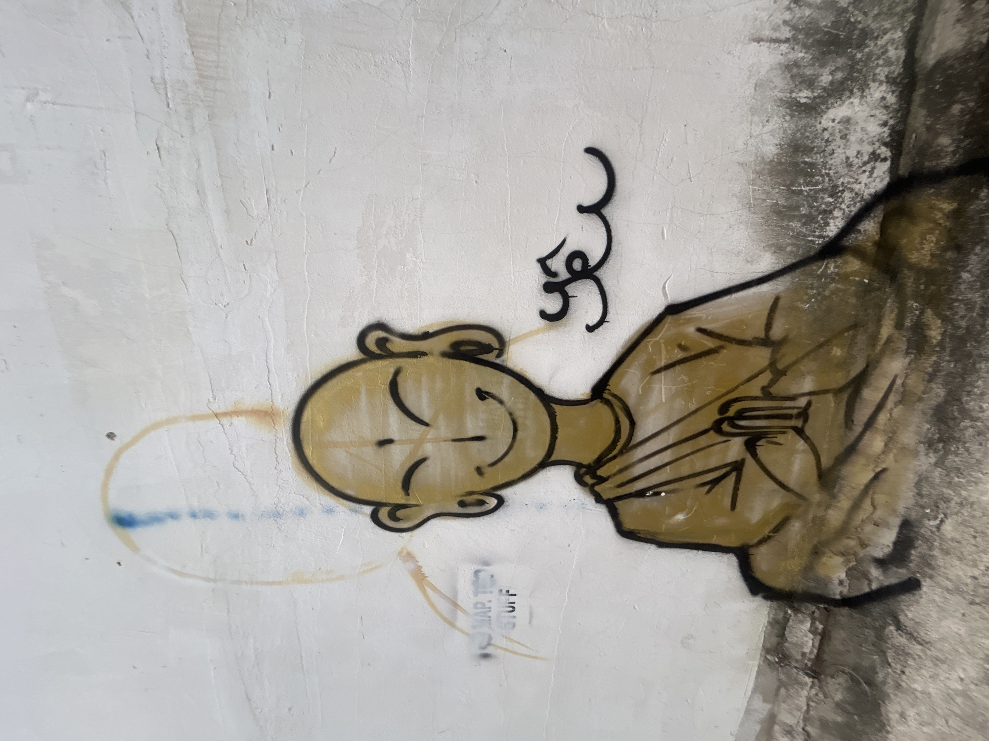 The image displays a piece of street art, a cartoon-style graffiti character, on a light-colored, textured wall. The main subject is a round-headed, bald figure with large ears, rendered in golden-brown tones with bold black outlines. The character has a serene expression, with simple lines forming closed eyes, a small nose, and a wide, closed-mouth smile. Its body is also golden-brown, clad in what appears to be a loose robe or tunic, with hands clasped together in front of its chest, suggesting a gesture of prayer or reverence.
Above the character's left shoulder (when viewed upright), a black, stylized symbol or script resembling a swirl is spray-painted onto the wall. To the right of the character's head, faint, circular, golden-brown spray paint marks are visible, along with a short, dashed blue line. The wall itself is uneven and rough, showing visible signs of wear, cracks, and some discoloration or grime, particularly on the right side where it transitions to a darker, mottled area.
Below the character's head, faint stenciled text is visible on the wall, which appears to read "MAP.TED STUFF". The overall impression of the artwork is one of peace and mild contemplation. The graffiti is situated in Bac Nha Trang Ward, Vietnam.