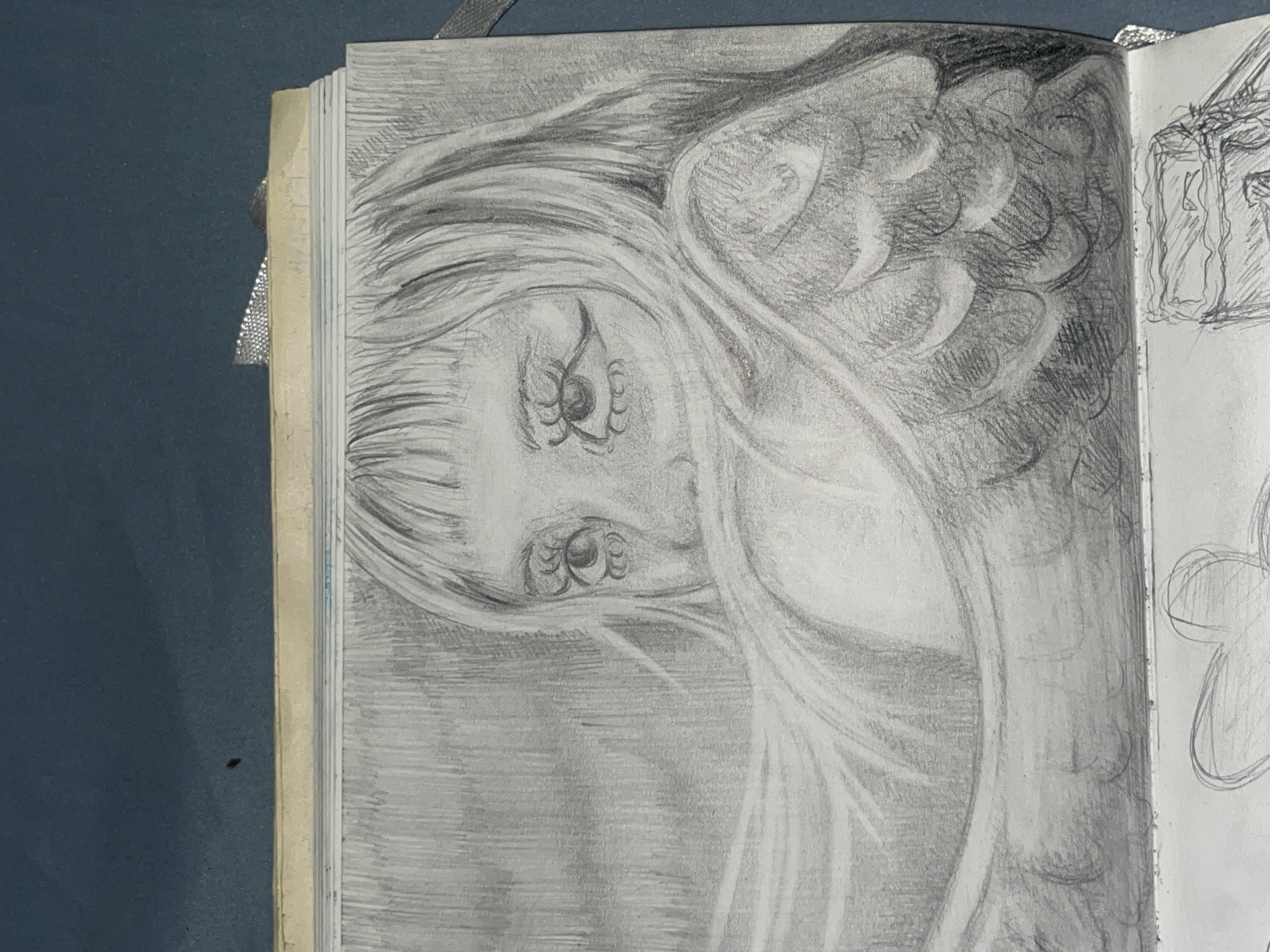 A black and white pencil sketch depicts a young woman's face partially obscured by her hair and what appears to be a wing. Her eyes are wide open with noticeable eyelashes and eyeliner. The sketch is on a page in a notebook, with a dark blue background visible above and to the right of the drawing. The notebook itself has a light yellow cover and white pages. The lower part of the image shows a glimpse of another sketch on the subsequent page, featuring two outlined hearts and a rectangular object with lines. The overall scene is indoors, with no indication of a specific city or location.