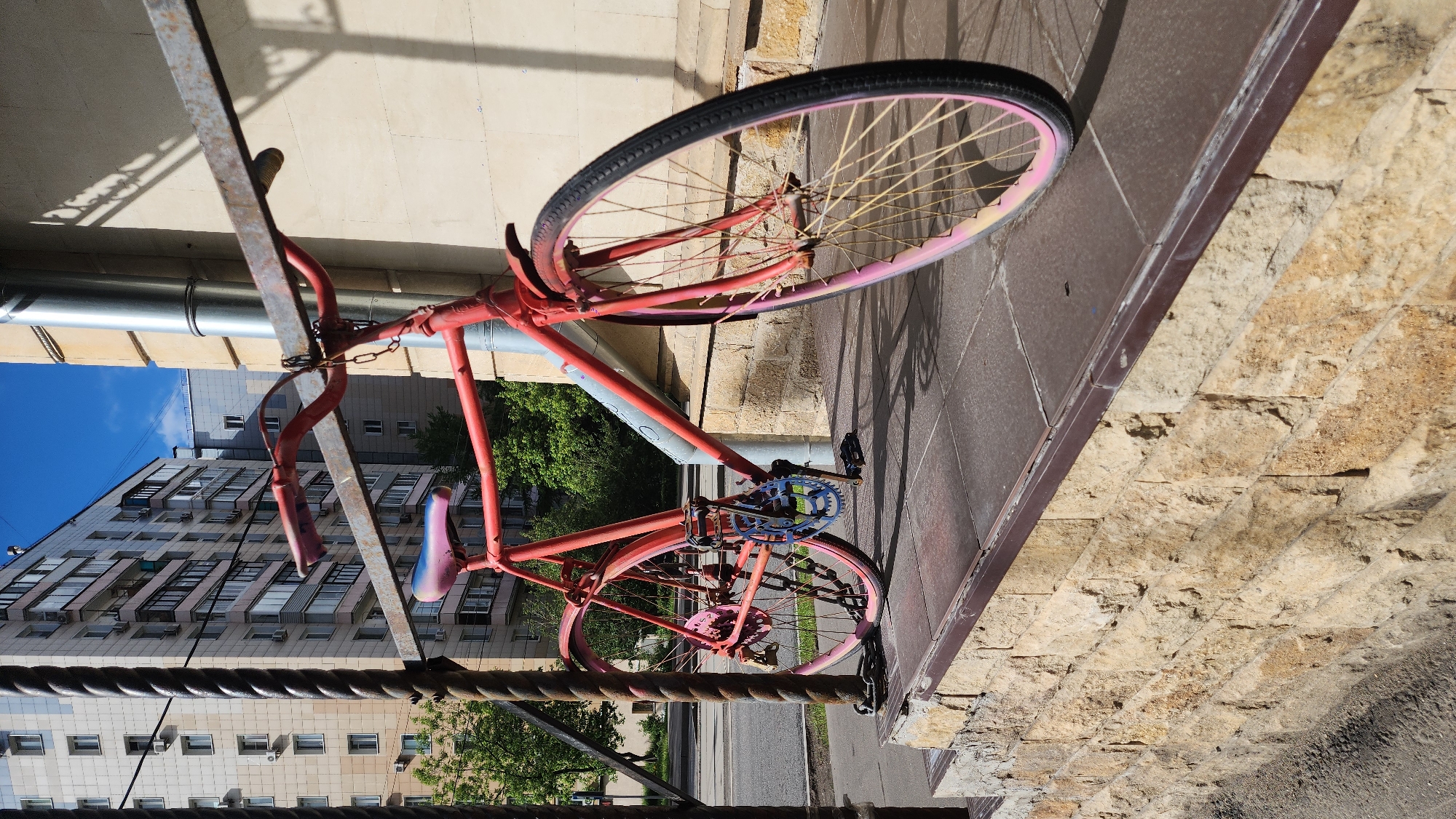 A red bicycle with pink rims is locked to a metal post in front of a building. The bicycle has a blue gear and a blue and pink seat. The building in the background appears to be residential. The location is likely in Moscow, Russia.