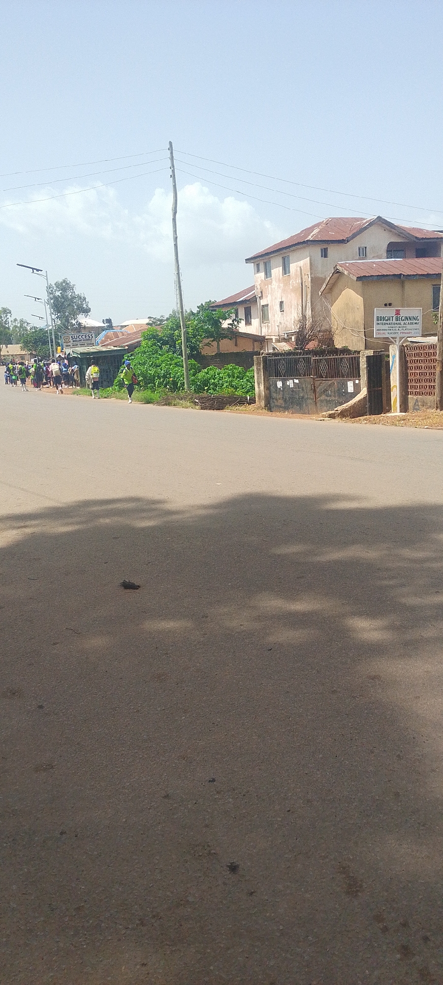 The image depicts a street scene with a group of people walking on the left side of the road. The people appear to be students, as they are dressed in matching uniforms and carrying bags. They are walking in the same direction, suggesting a procession or a school activity. The road is paved and appears to be in an urban or semi-urban environment.
On the right side of the road, there are buildings. One building has a sign that reads "BRIGHT BEGINNING INTERNATIONAL ACADEMY". This suggests that the location might be near a school. The buildings have a somewhat aged appearance with red-tiled roofs. There is a gate in front of one of the buildings, and some vegetation is growing along the street.
In the foreground, the asphalt road is visible, with shadows of trees cast upon it. The sky is mostly clear with some scattered clouds. A wooden utility pole with wires is also present in the scene. The overall impression is a casual daytime outdoor activity on a street in a populated area.