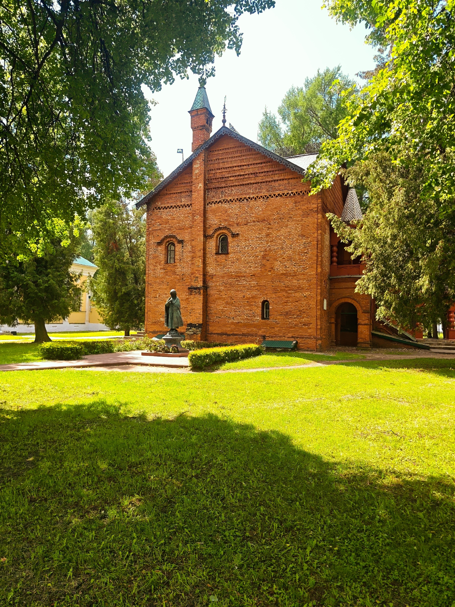 The image depicts a red brick building in a grassy area with trees. A statue stands in front of the building. The location is Unknown, Unknown.