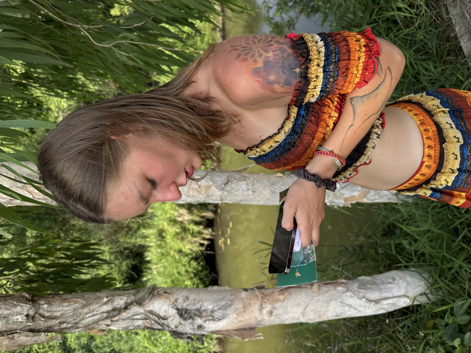 A young woman with shoulder-length brown hair and fair skin, wearing a brightly colored, off-the-shoulder top and skirt, stands outdoors near a body of water and trees. She is holding a smartphone and a green booklet in her hands, and appears to be looking at the phone. She has tattoos on her right shoulder and arm, and is wearing bracelets on her left wrist. The environment suggests a natural setting, possibly a park or rural area, with lush green vegetation and a calm body of water in the background. The presence of trees and water implies an outdoor location, possibly in a tropical or sub-tropical climate. Without further information or context specific to Nam Hoa Lư Ward, Vietnam, pinpointing the exact location within that city is not possible based solely on the visual information in the image.