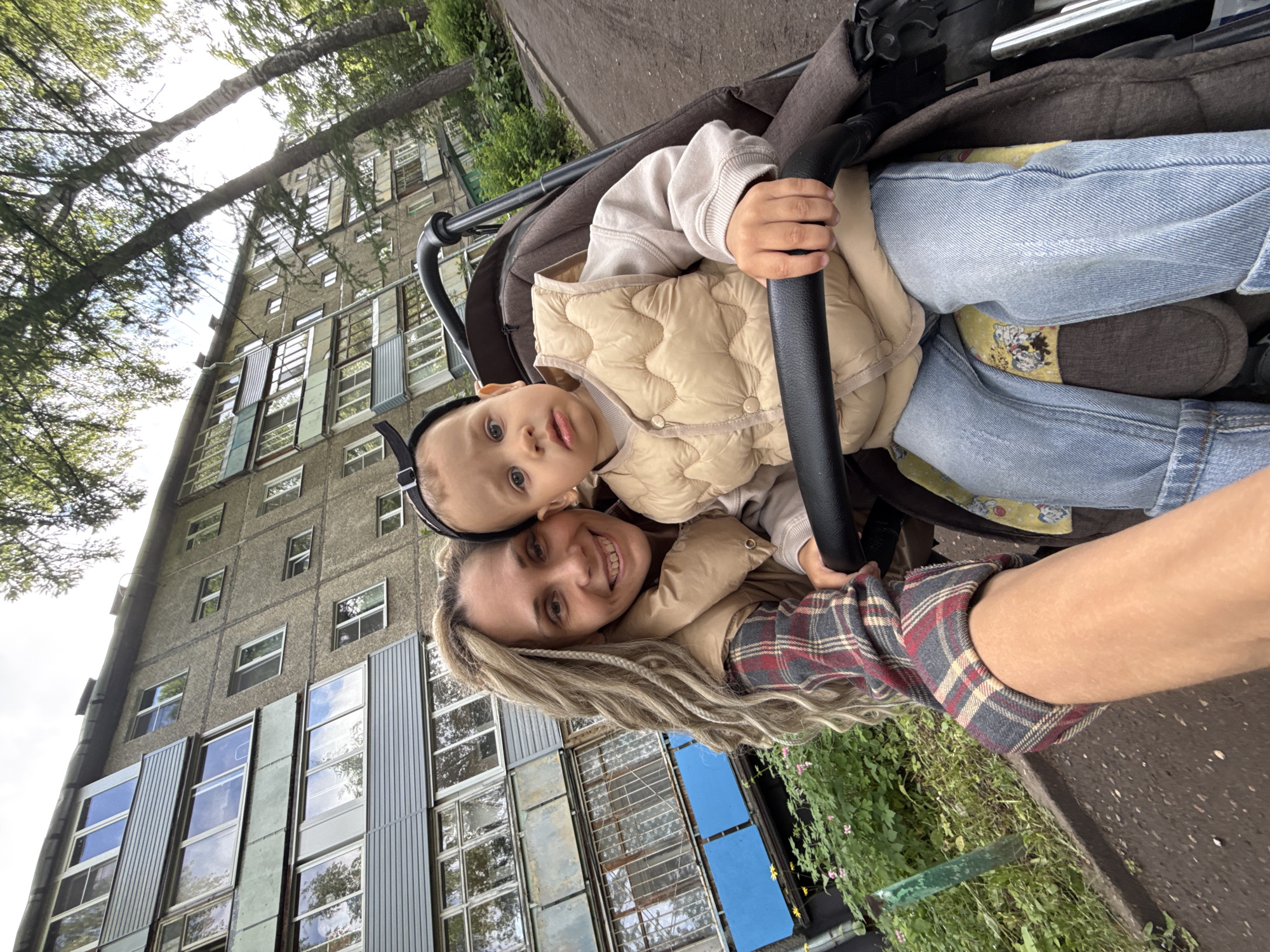 A mother and child pose for a selfie in Rusanovo, Russia. The child, wearing a beige vest and blue jeans, sits in a stroller and holds onto the stroller's bar. The mother, with blonde braided hair and wearing a plaid shirt, smiles at the camera. Behind them is a tall, grey building with numerous windows and blue metal panels. Green foliage is visible nearby, and the ground appears to be paved.
