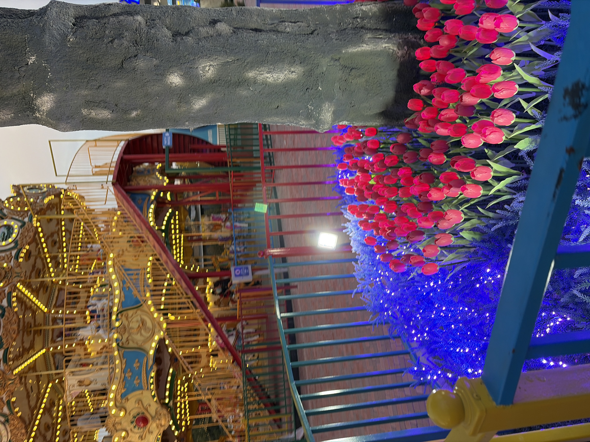 The image depicts a brightly lit carousel at an amusement park, with a large tree trunk in the foreground. The carousel is yellow and blue, adorned with numerous lights, and features a prominent red staircase leading up to its levels. In front of the carousel, a fence separates it from a garden bed filled with red tulips and accented by blue lights. The sky above is overcast, suggesting it might be evening or dusk. The scene is set in an outdoor urban environment, likely an amusement park.