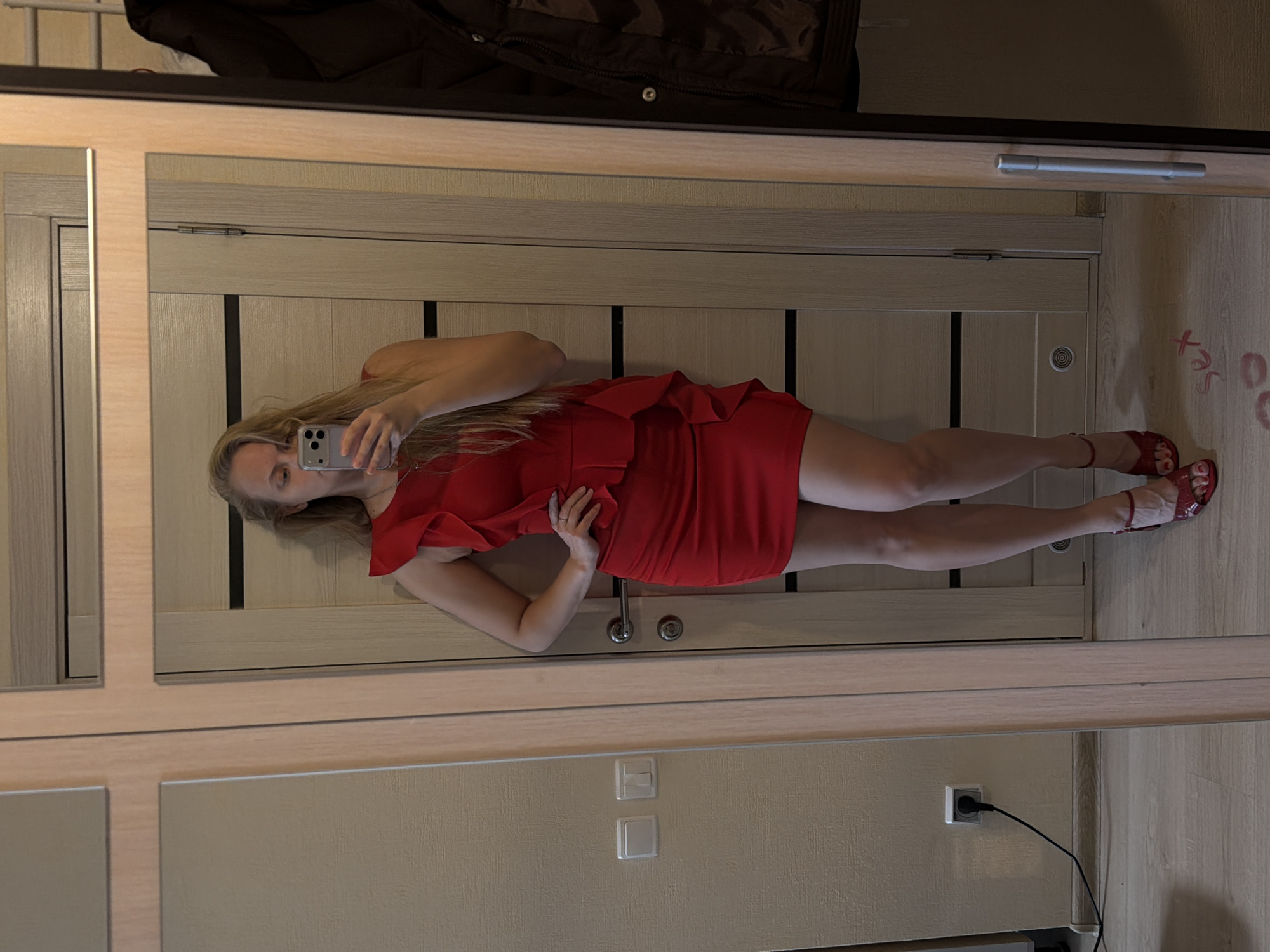 A woman is reflected in a mirror. She is holding a smartphone and wearing a red dress and red sandals. The reflection shows her standing in what appears to be an interior space with light-colored furniture and a wooden door. The room has electrical outlets and switches. The location in Izhevsk, Russia, is not apparent from the image.