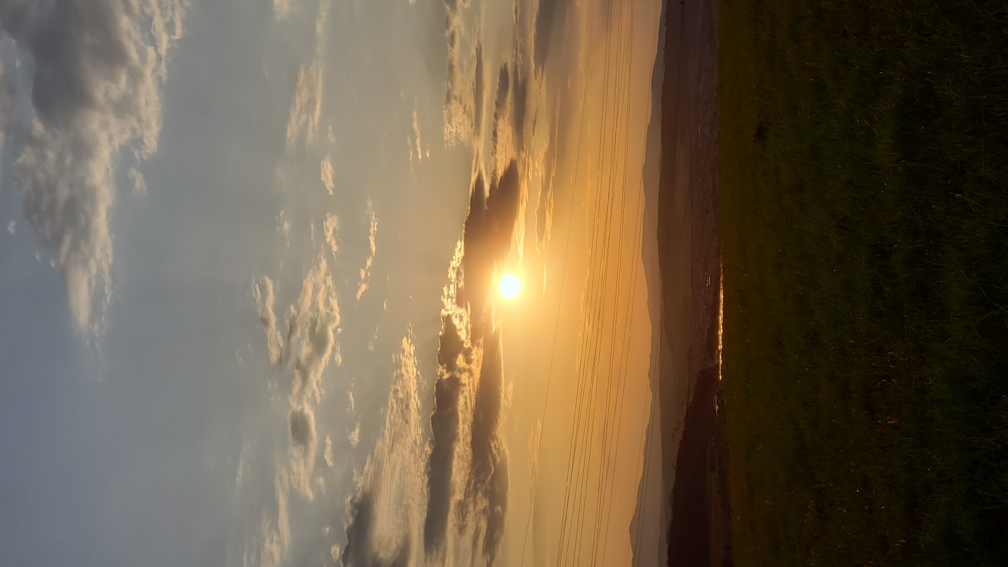 The image depicts a sunset over a rural landscape, possibly near the city of Prešov, Slovakia. The sun, a bright orange orb, is partially obscured by clouds, casting a warm glow across the sky. Rays of light emanate from the sun, creating a dramatic effect. In the foreground, a grassy field is visible, with a line of trees and hills in the distance. A body of water, possibly a river or lake, reflects the golden light of the sunset. Power lines stretch across the horizon, a subtle reminder of human presence in the otherwise natural setting. The overall atmosphere is one of tranquility and natural beauty.