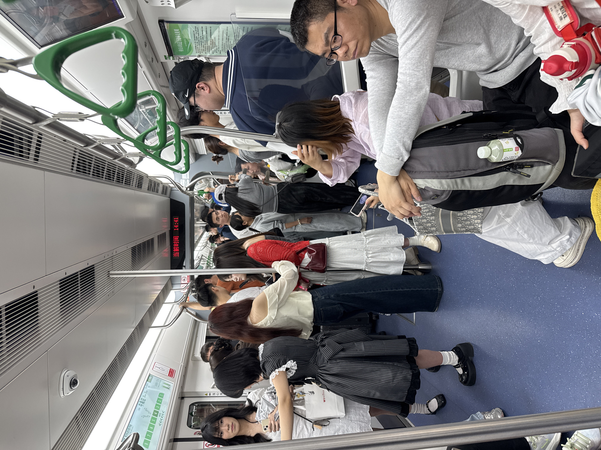 Inside a subway car in Yuzhong District, China, a diverse group of people are standing. The subway car has green handrails. A digital display shows the current time as 14:41. One advertisement for "WINONA" shows up in one window. One screen shows the Yuzhong District train line.