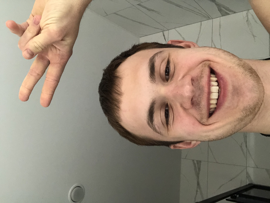 The image features a young adult male, likely taking a selfie or being photographed, with a wide, joyful smile. The image is rotated 90 degrees clockwise, so if viewed upright, the man's head would be on the left side of the frame, looking slightly upward and to the left, with his hand positioned above and to the left of his head.
The man has short, dark brown hair and light stubble covering his chin and jawline. His face is expressive, dominated by a broad smile that reveals his upper and lower teeth. His eyes are dark brown and appear bright and cheerful. He has well-defined eyebrows.
In the upper left portion of the image (or above his head if upright), a fair-skinned hand is visible, making a "V" sign or peace sign. The index and middle fingers are extended, while the thumb, ring finger, and pinky finger are curled into the palm. The skin on the hand shows some texture.
The background consists of two main sections. To the right of the man's head (bottom right if upright), there's a wall with white and light grey marble-like tiles, suggesting an indoor setting like a bathroom or kitchen. The rest of the background is a plain, light grey wall or ceiling surface. In the bottom left corner of the original image (which would be near the bottom of the plain grey wall if upright), a circular, light-colored object, possibly a light fixture or vent, is visible. A dark, angular object, possibly part of furniture or a frame, is partially visible in the very bottom right corner of the original image (bottom edge of the tiled wall if upright).
The overall mood of the image is positive and lighthearted, conveyed by the man's wide smile and cheerful expression. There is no visible text in the image.