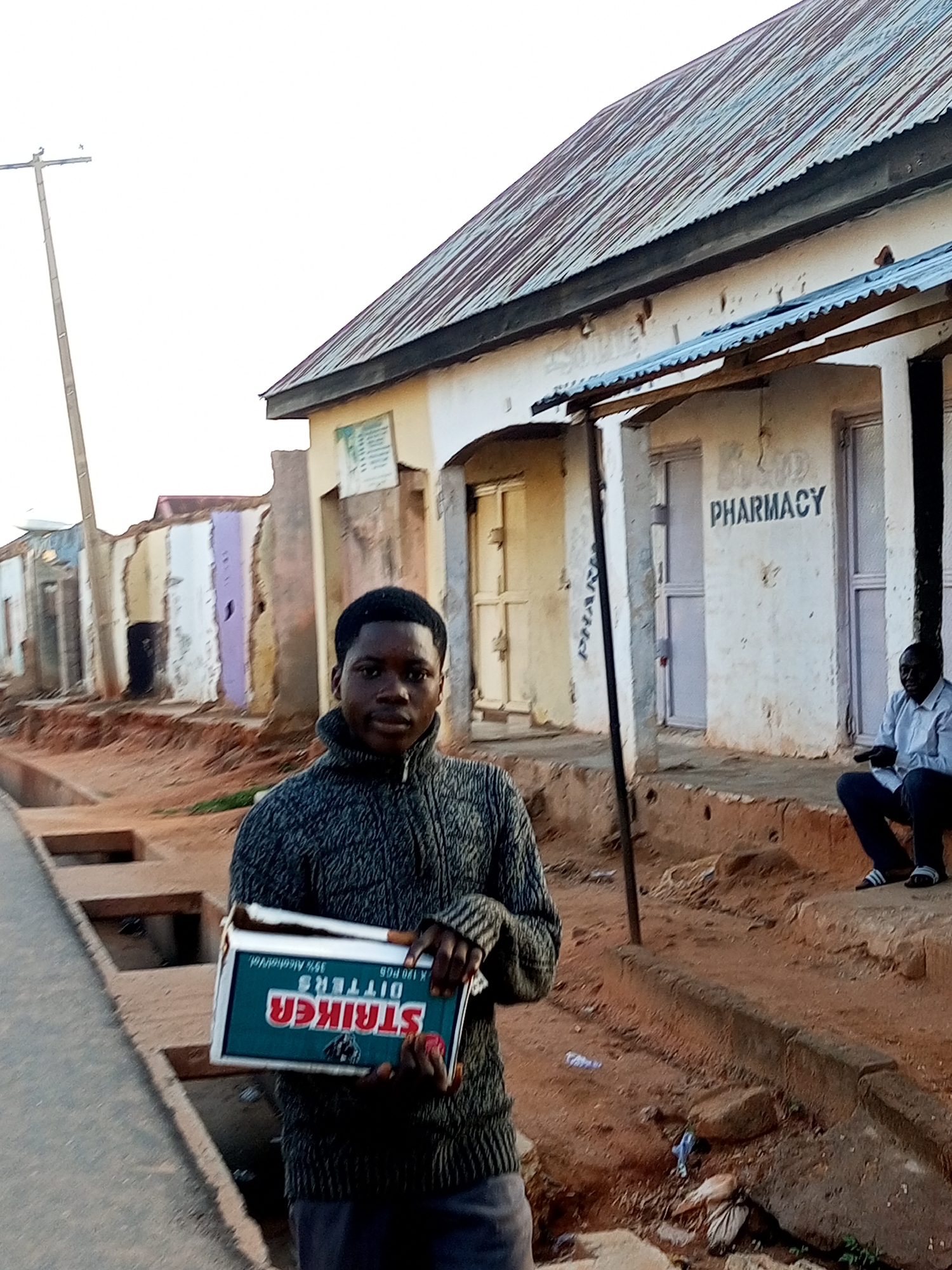 In Gombe, Nigeria, a young man stands on a dirt road next to a dilapidated building that appears to be a pharmacy. He is holding an open box of what seems to be cigarettes. The building has a corrugated metal roof and faded paint. Behind him, other structures show signs of disrepair. A man in a white shirt and dark pants is seated on a step in front of the pharmacy, looking towards the camera. The sky is overcast, suggesting either early morning or late afternoon.