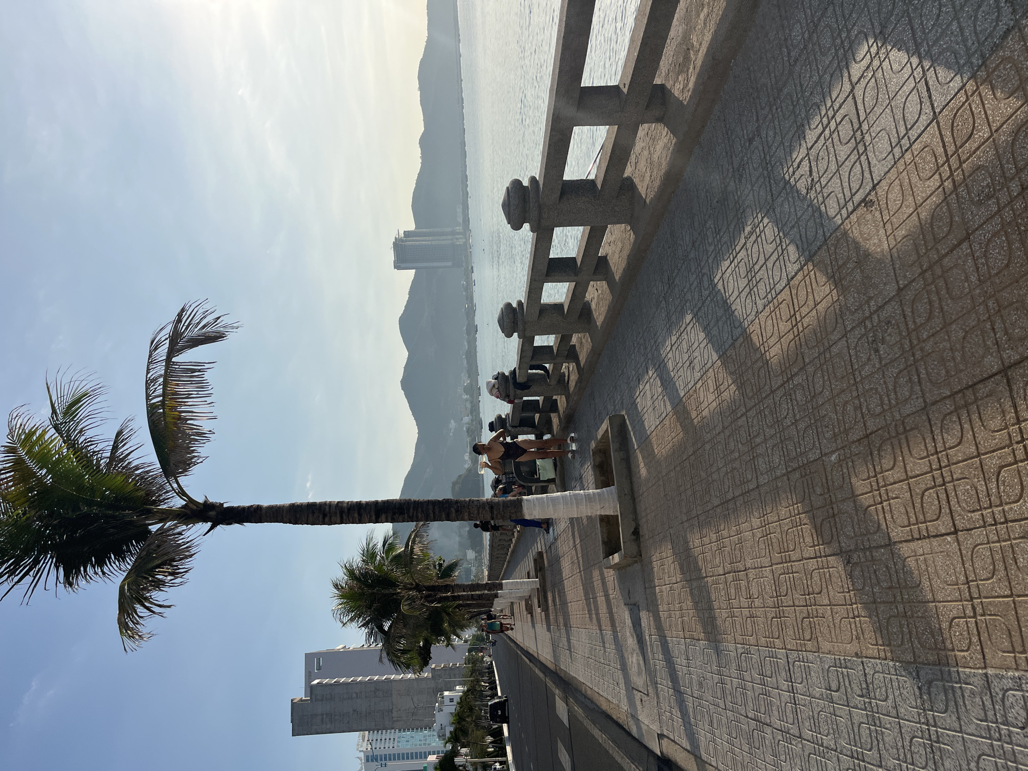 The image depicts a coastal scene in Bac Nha Trang Ward, Vietnam. A tiled sidewalk runs along a concrete barrier, casting shadows on the ground. Beyond the barrier is the sea, with mountains visible in the distance and buildings along the coastline. Palm trees are prominently featured, one to the left and another partially obscured by the first. People are standing near the barrier. A person dressed in black stands by the wall drinking water. In the distance are other people walking along the seawall. A tall building is visible near the palm tree. The sky is bright and hazy. A street runs parallel to the sidewalk.