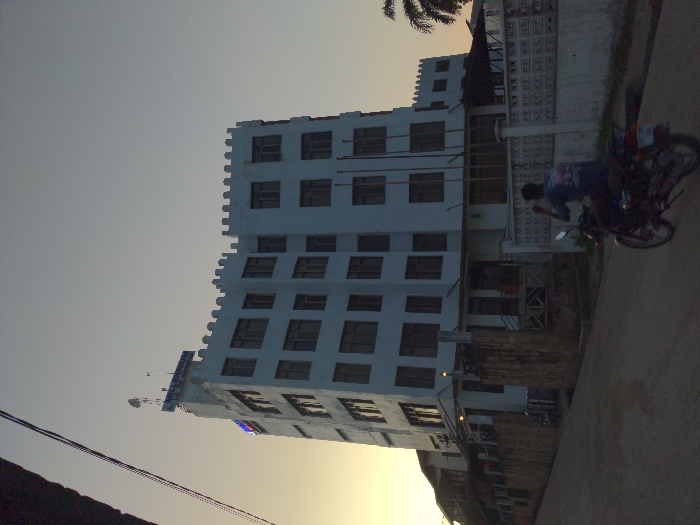 This image depicts a street scene in Bagamoyo, Tanzania, likely during the late afternoon or early evening. The sky, which dominates the upper left, transitions from a light grey-blue to a warm, golden hue near the horizon, indicative of sunset.
The central subject is a tall, multi-story white building characterized by a distinctive castellated parapet along its roofline, giving it a castle-like aesthetic. Its facade is punctuated by numerous rectangular windows. On the upper left side of the building, a blue sign is visible, with a portion of the text appearing to read "BAGAMOYO," a clear location cue. Another smaller blue sign is located further down the building's side.
In the foreground and to the right, a paved street runs alongside the building. A person is seated on a motorcycle on the street, facing away from the camera. A decorative, white patterned wall or fence separates the building's property from the street. The top right corner shows a glimpse of palm tree fronds. The scene appears calm and everyday, with the warm light casting a soft glow over the surroundings.