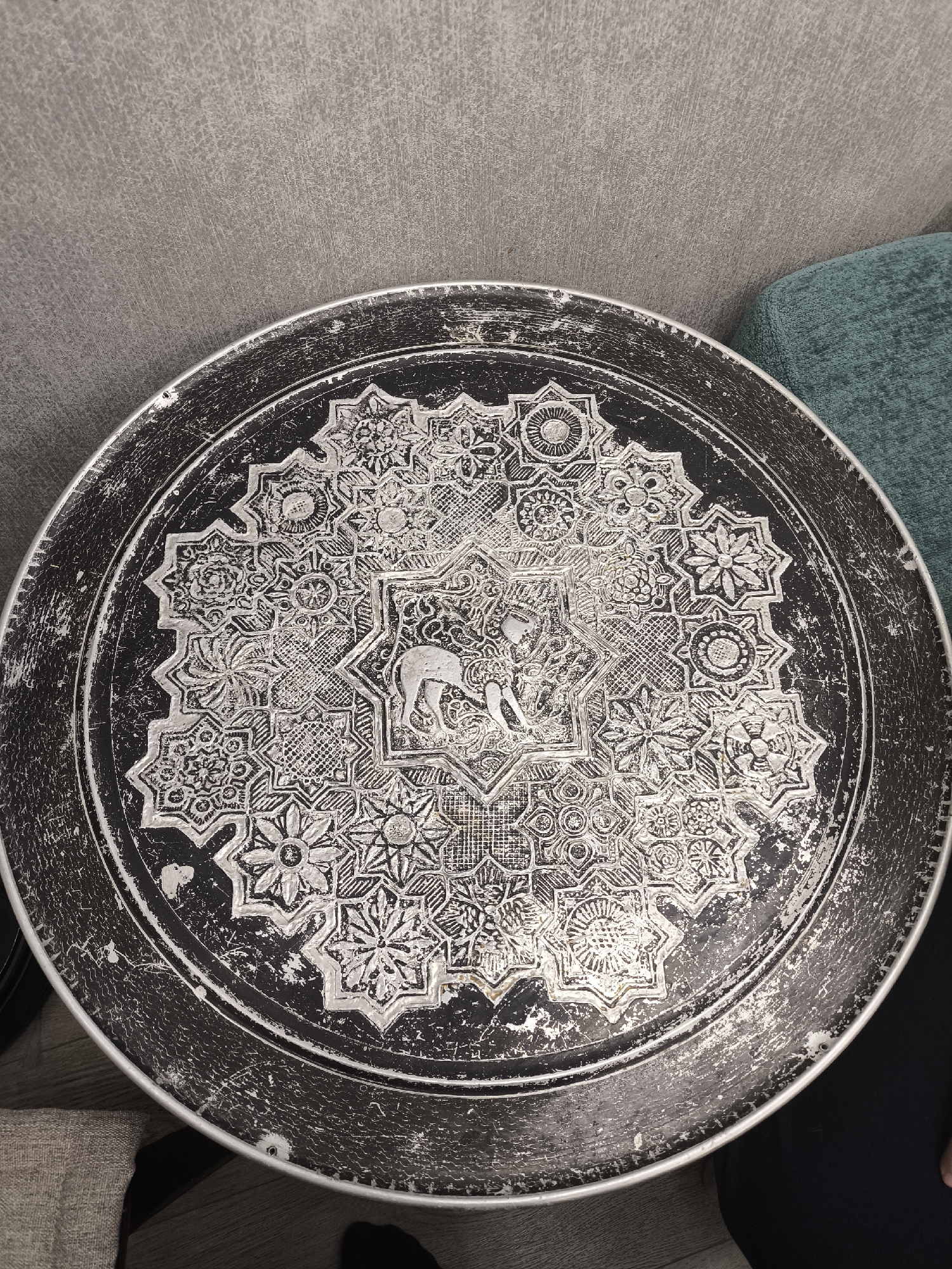 The image shows a round, possibly metal, tray or table top with intricate geometric and floral designs etched into its surface. A stylized image of a camel is centered within a star-shaped section of the design. The overall aesthetic appears to be of Middle Eastern or North African origin. The tray is placed on or near a surface, with a portion of a green fabric item (possibly a cushion or part of a chair) visible in the upper right corner and a neutral colored item on the lower left. The surrounding environment appears to be indoors, judging by the presence of upholstered furniture. No people are visible in the image. There is no indication of the location being Moscow, Russia.
