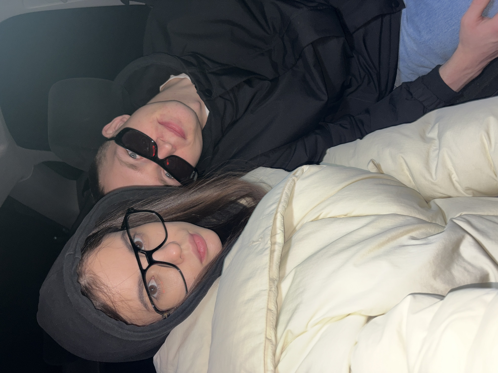 The image displays two young individuals, one male and one female, lying down in what appears to be the interior of a vehicle, likely resting or relaxing. The scene is illuminated by dim, artificial light, suggesting either an indoor setting or nighttime.
The male subject, positioned towards the top left, wears a black jacket over a light-colored shirt and dark, rectangular sunglasses. He looks directly at the camera with a neutral expression and slightly parted lips. Behind him, a dark car seat or headrest is visible.
The female subject, situated towards the bottom right, wears a black hooded top and large-framed glasses. She also looks towards the camera with a calm expression, her long brown hair framing her face.
Both individuals are partially covered by a light-colored, possibly cream or off-white, quilted blanket or duvet, which suggests a cozy and comfortable environment. The blanket occupies a significant portion of the right side of the frame, showing its soft, textured surface. The overall mood of the image is relaxed and peaceful. The image is rotated 90 degrees clockwise. No text is visible in the media file. The context provided for the location is Chaikovsky, Russia.