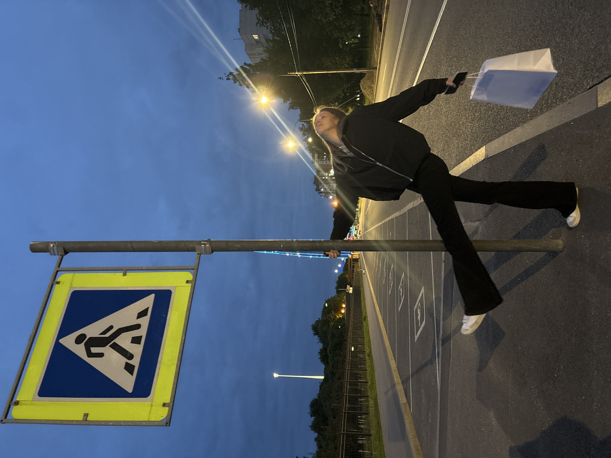 A woman is standing on the edge of a sidewalk next to a road in Saint Petersburg, Russia, seemingly posing for a picture. She is positioned sideways, holding onto a metal pole, part of a pedestrian crossing sign. The sign itself features a blue square with a white triangle, inside which is a black figure indicating a person walking. The sign is framed in bright yellow.
The woman is wearing a dark jacket and black trousers, with white sneakers. She is holding a white shopping bag in one hand. The background shows a road with parking spaces, trees, street lights, and buildings. The sky is a dusky blue, suggesting it is late evening or early morning. The lighting in the photo is a mix of natural twilight and artificial street lighting.