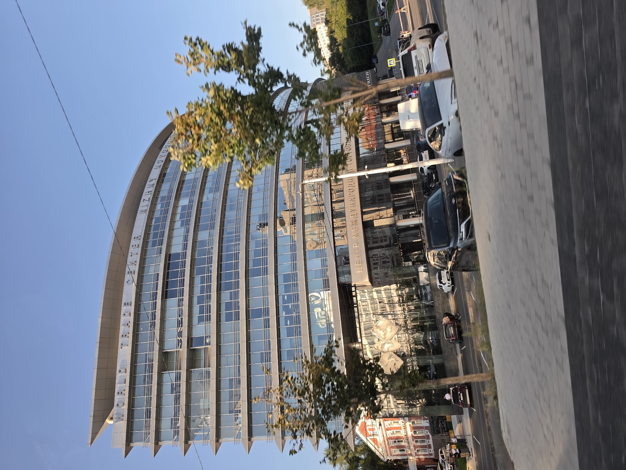 The image shows the World Trade Center building in Nizhny Novgorod, Russia, on a clear, sunny day. The modern, curved glass building is the dominant feature, with "WORLD TRADE CENTER" visible across its upper facade. Below the main structure, the building's glass facade reflects the surroundings, including trees and other buildings.
In front of the World Trade Center, a street is visible with several cars parked or driving. Among them, a white sedan, a dark SUV, and a black sedan are clearly identifiable. There are also a few individuals walking on the sidewalk or near the cars. To the left of the World Trade Center, a red brick building with white accents is partially visible. Lush green trees, some with yellowing leaves indicating autumn, are scattered throughout the scene. The sky is a bright, clear blue. The perspective suggests the photo was taken from a moving vehicle.
