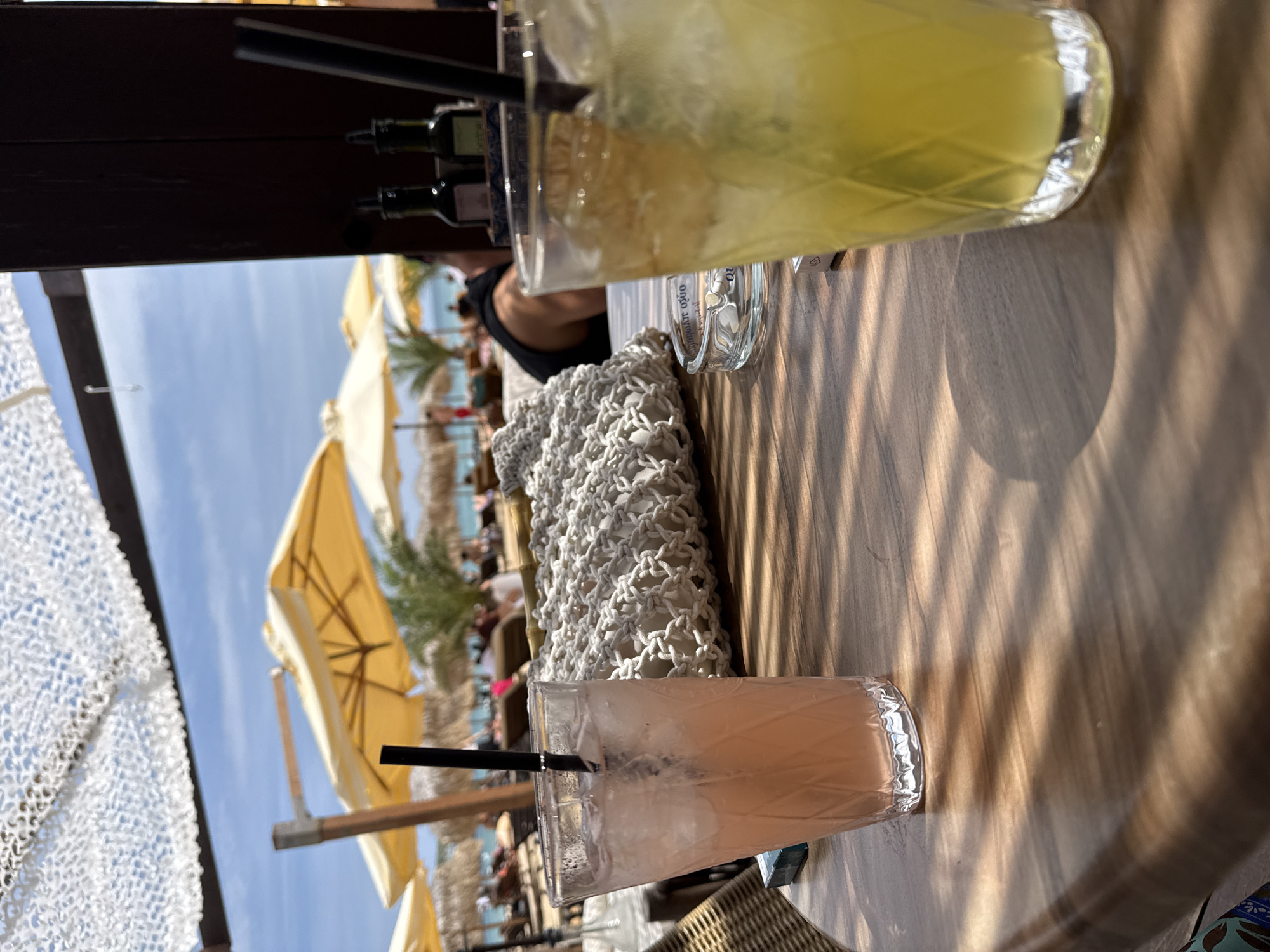In an outdoor beach setting, a close-up shot focuses on a wooden table with two drinks and a woven bag. The drink on the left is a pink cocktail with ice and a black straw, served in a patterned glass. To its right, a yellow-green cocktail with ice and a garnish is in a similar glass. A white woven bag sits behind the drinks. An ashtray with cigarette butts and a pack of cigarettes are visible to the left of the bag. In the background, yellow umbrellas, thatched-roof beach huts, and palm trees line a sandy beach with people lounging and the ocean beyond. The sky is blue with scattered clouds, and a white canopy is overhead. The scene suggests a relaxed atmosphere at a beach resort.