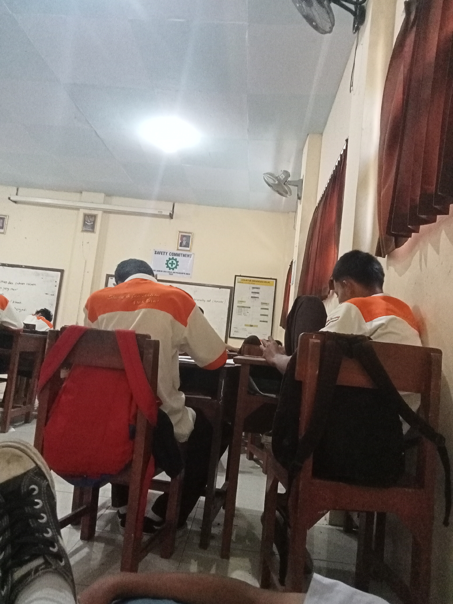 The image depicts an indoor scene, likely a classroom in Secang, Indonesia. In the foreground, a person's legs and feet are visible, wearing black Converse sneakers, suggesting they are sitting or lying down. The background shows several students seated at wooden desks, facing forward. One student in the middle ground wears an orange and white shirt, with text on the back. Another student to the right, also wearing a similar shirt, is seated and appears to be engaged with their phone. The classroom environment includes a whiteboard with writing, posters on the wall, and a ceiling fan. Red curtains are visible on the right side of the image. The lighting suggests it's daytime, with artificial lights on the ceiling also contributing to illumination.