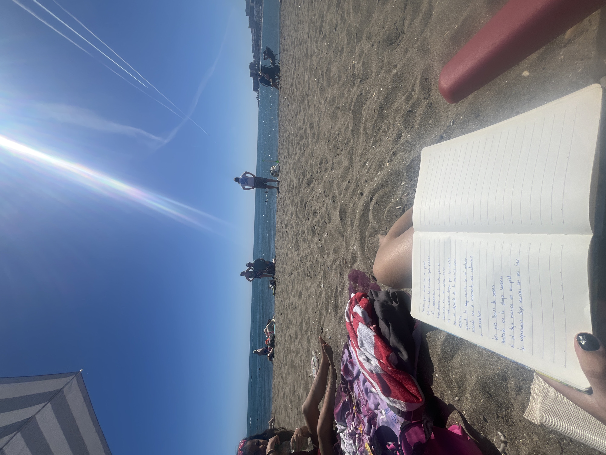 The image depicts a person sitting on a beach with a notebook in their lap, writing in it. The beach is sandy, and there are other people in the background. A blue sky with plane contrails overhead. The location is Arroyo de la Miel-Benalmádena Costa, Spain.