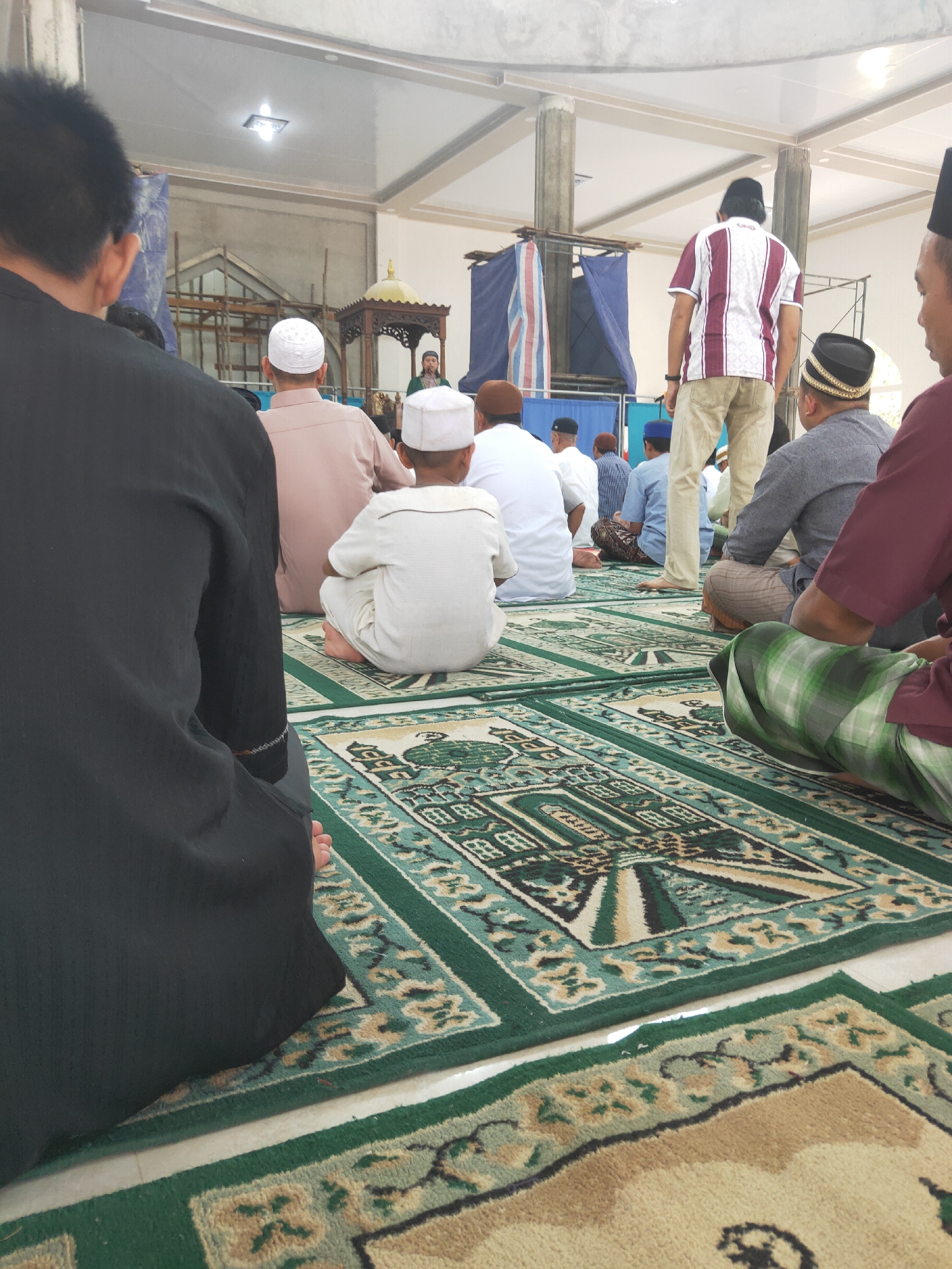 In Sintang, Indonesia, a group of men are seated on patterned prayer rugs inside a building, likely a mosque. The men are facing the front of the room, where a speaker is visible, giving a sermon. The room has a high ceiling with visible support beams and lighting fixtures. Construction appears to be ongoing in the background, with scaffolding and blue tarps. The men are dressed in traditional Islamic attire, with many wearing kufis and long shirts or robes. Some are in the process of prayer. The overall atmosphere suggests a religious gathering or service.