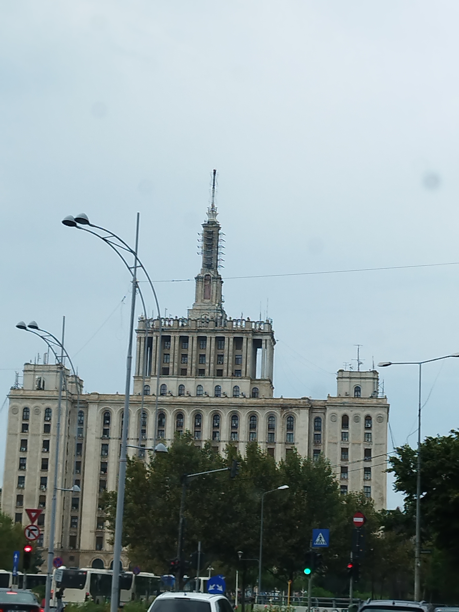 This is a full shot of the Palace of the Parliament building in Bucharest, Romania, captured on a cloudy day. The massive Brutalist structure dominates the frame, characterized by its imposing scale, symmetrical design, and numerous windows. The building is adorned with architectural details such as columns and a prominent central spire. In the foreground, a busy street scene unfolds with multiple vehicles, including cars and buses, on the road. Traffic lights and street lamps are visible, indicating an urban setting. Lush green trees line the street, providing a contrast to the concrete of the building. The overall impression is one of an overcast day in a bustling metropolitan area.